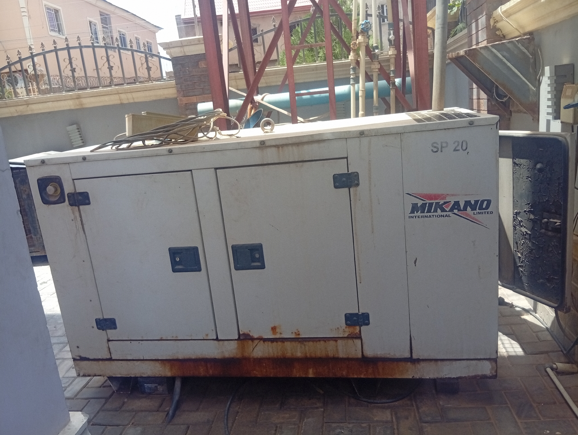 The image features a Mikano SP 20 generator, displaying signs of rust along its base. The generator is situated in Aviation Quarters, Nigeria. In the background, there's a house with an iron fence and red metal support beams. There are also visible pipes and some vegetation in the background. The generator is on a brick-paved surface.