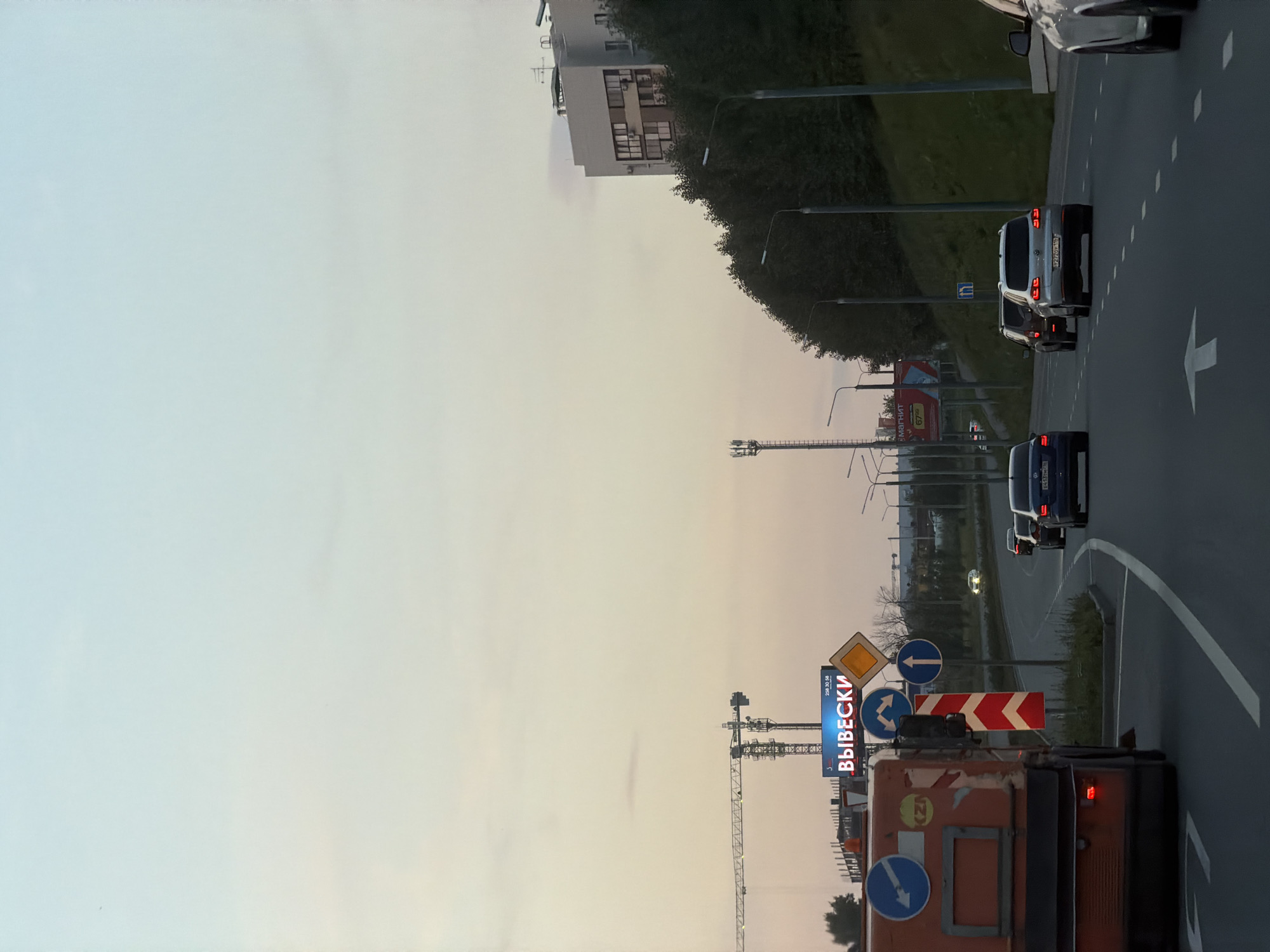 The image shows a street scene at dusk, possibly in a suburban or industrial area. In the foreground on the left is the rear of an orange vehicle, possibly a sanitation truck, with various signs and markings on its side. To the right of this vehicle, a road curves to the right, with several cars driving in the same direction. In the background, there are trees and a building on the right side of the road, and on the left side, there is a billboard with the word "ВЫВЕСКИ" (meaning "Signs" in Russian) illuminated in blue, as well as construction cranes. The sky is a pale gradient of blue and pink, indicating the time of day is around sunset or sunrise. There are streetlights visible along the side of the road.