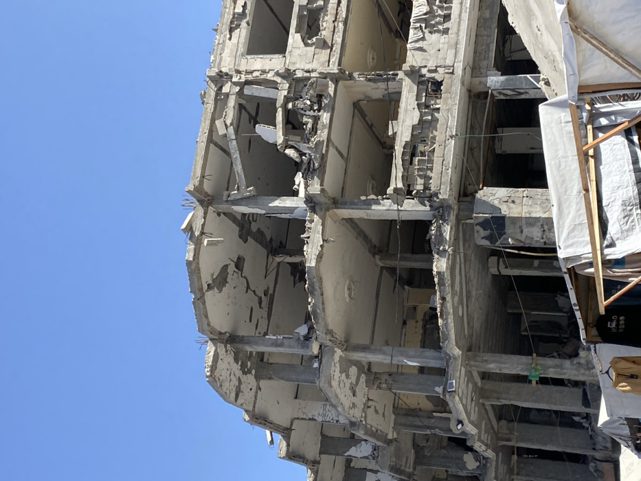 The image displays a multi-story concrete building in Khan Yunis, Palestinian Territory, exhibiting severe structural damage under a clear blue sky. The building's upper and middle sections are extensively compromised, with multiple floors partially collapsed, walls largely absent, and internal concrete columns, beams, and slabs exposed. Fractured concrete, twisted rebar, and debris are visible across the remaining floor plates and ledges. In the lower foreground, several temporary structures, appearing as makeshift shelters or stalls, are constructed from wooden frames and covered with white and light-colored tarpaulins or fabrics. Various items of clothing, including a yellow jacket and a black shirt with white text, are suspended from these temporary constructions. No individuals are visible within the frame. The scene is illuminated by natural daylight.