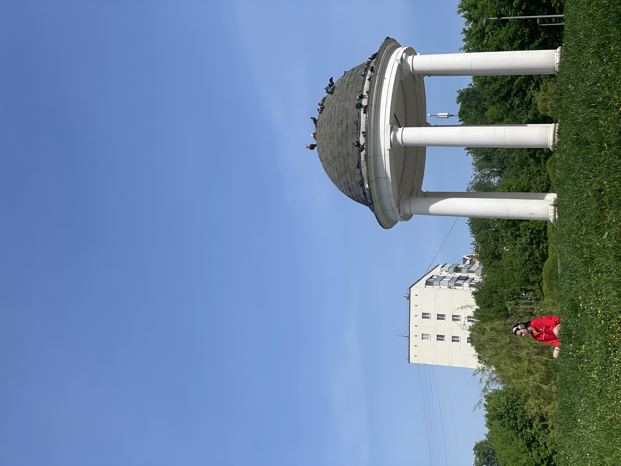 The image shows a rotunda with pigeons on the dome, located in a park in Moscow, Russia. The sky is blue, and the rotunda is supported by white columns. A person wearing a red jacket and headphones is sitting in the grass nearby. A white building with balconies is visible in the background. There are trees and green vegetation throughout the scene.
