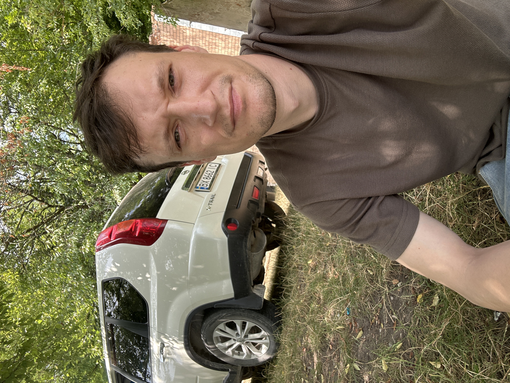 The image is a selfie taken outdoors, likely in or near Makiv, Ukraine. In the foreground, a man with short brown hair and a light beard is partially visible, wearing a brown t-shirt and blue jeans. He is looking towards the camera with a serious expression. Behind him, a white SUV is parked on a grassy area. The vehicle has a license plate that reads "BX 8662 IC". The background consists of lush green trees and a section of a brick wall. The overall impression is a casual outdoor setting on a sunny day.