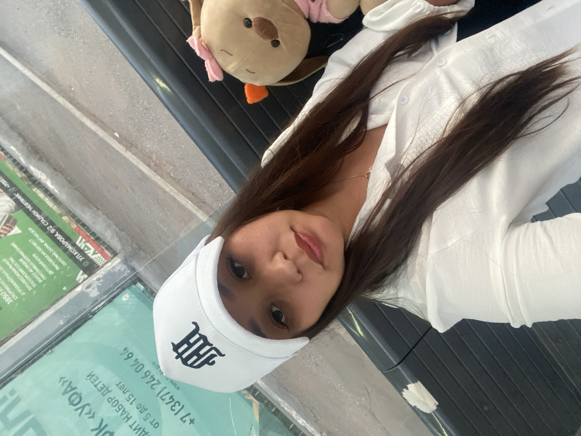 The image shows a young woman wearing a white baseball cap with a black logo and a white button-up shirt. She is sitting next to a plush toy with a pink bow. In the background, a green sign with Russian text is visible. The sign contains the text "УК «УФА»" and "СПОРТ.", indicating that the location is likely in Ufa, Russia, and related to sports. Additional text on the sign appears to be advertising or informational, including phone numbers and a website. The environment appears to be outdoors, possibly near a transportation hub or public space, given the presence of seating and the background signage. The woman is looking towards the camera, and the overall impression is a casual, self-taken photograph.