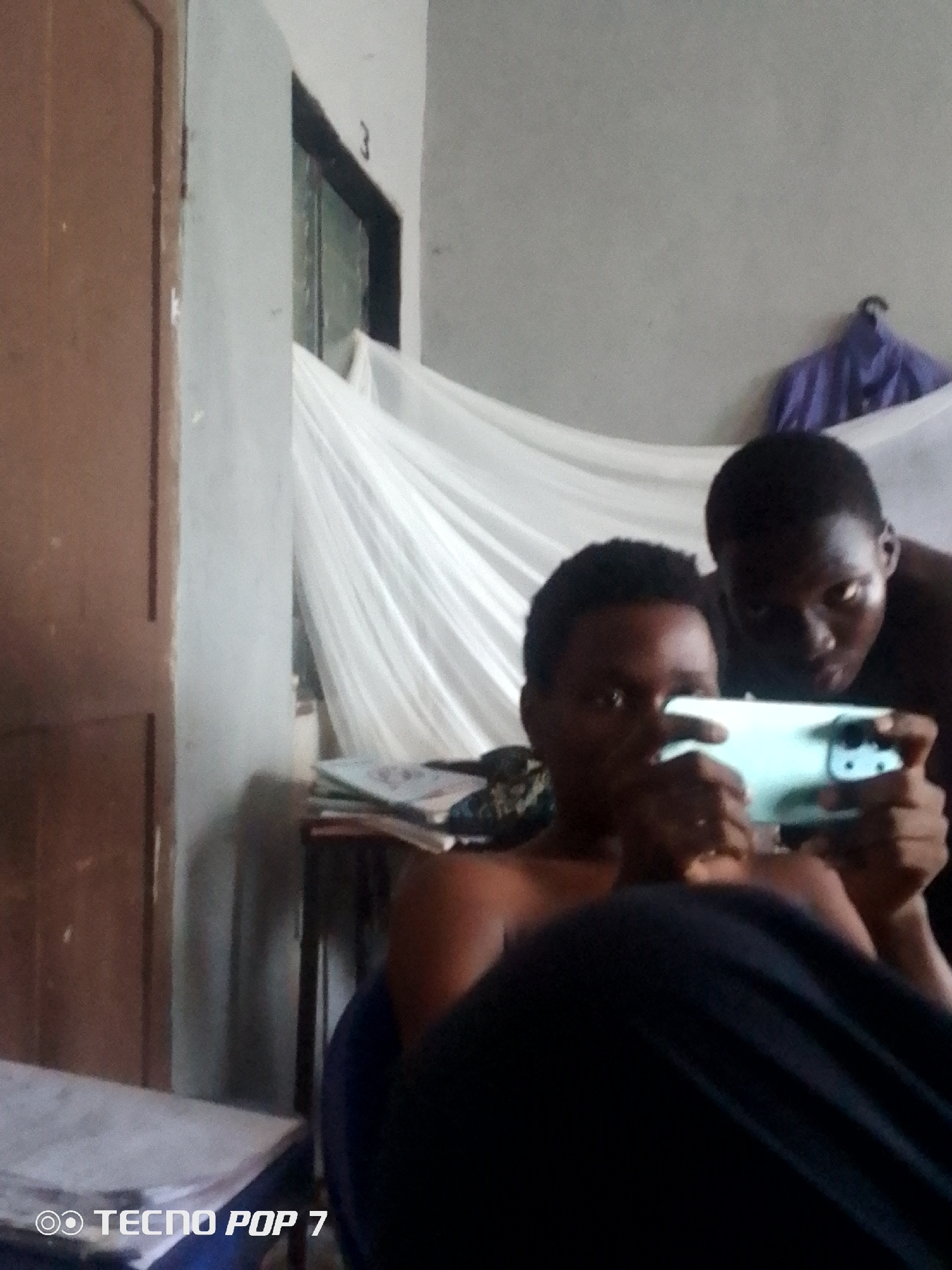 The image shows two young men in what appears to be a bedroom in Ipetumodu, Nigeria. The room has a wooden door on the left, a window with a mosquito net draped over it in the background, and a pile of books or papers on a shelf or desk to the right. A purple garment is hanging on the wall behind the men. One young man in the foreground is holding a light blue smartphone in landscape orientation, appearing to be playing a game or watching something on it. He is shirtless, as is the other young man standing behind him, who is looking at the phone. The lighting in the room is dim, suggesting it might be daytime with diffused light or evening. The overall impression is of a casual, indoor scene within a residential setting in Nigeria.