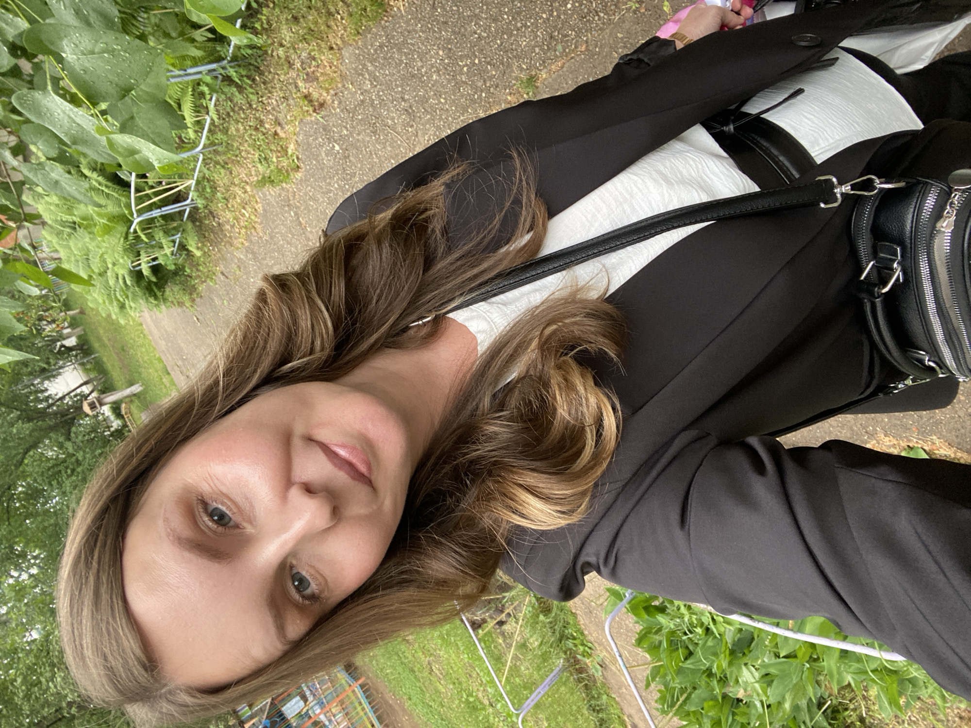 A woman is facing the camera at an angle, with a background of greenery and a pathway. She has medium-length, light brown hair with highlights and is wearing a black blazer over a white top. She has fair skin, visible eyelashes, and light-colored eyes. A black handbag is slung over her shoulder. The background includes a pathway, a small patch of grass, and various plants and shrubbery. There's a small blue and red play structure with a swing set. The specific location is unclear from the image.