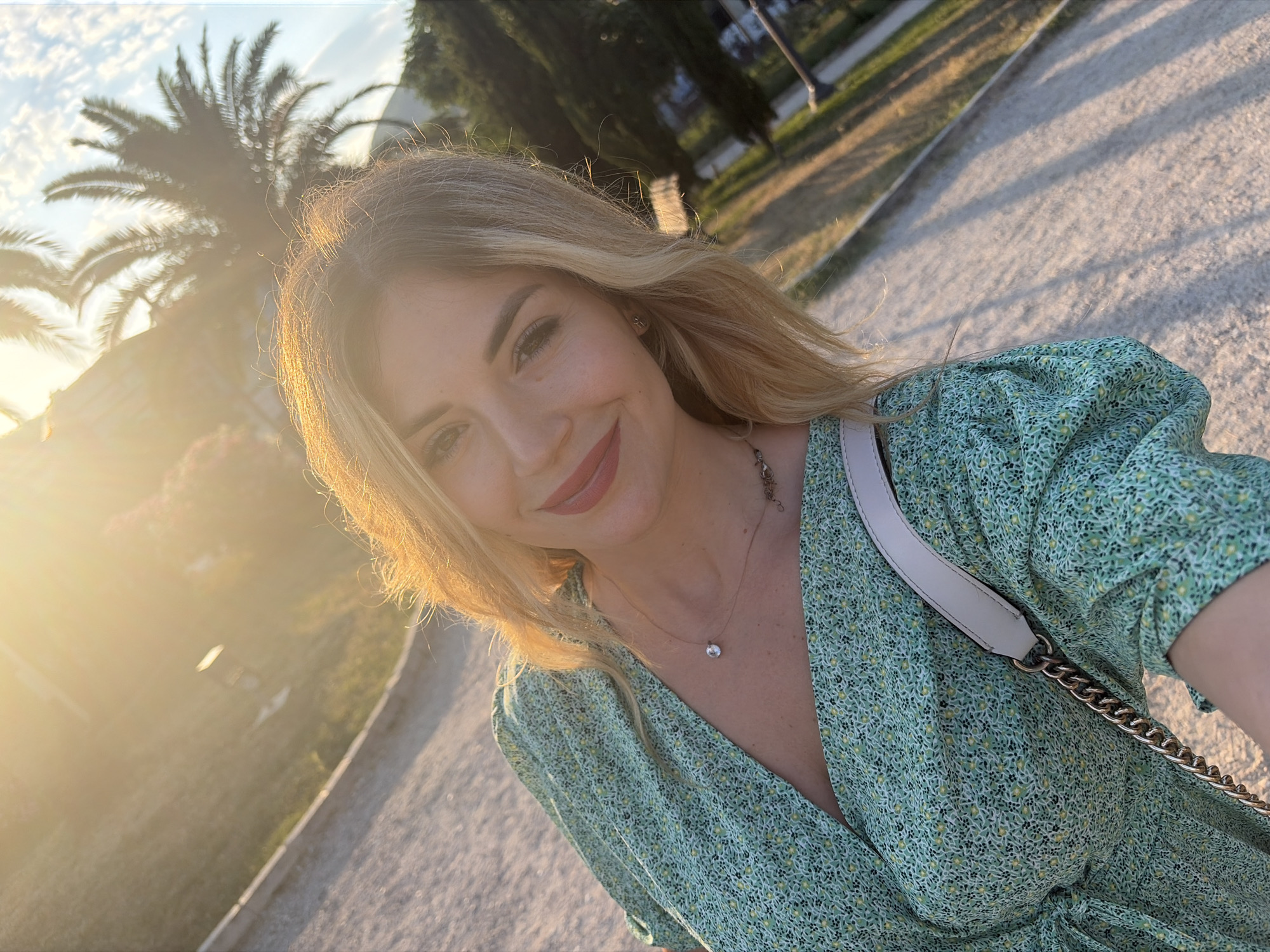 This media file is a close-up selfie of a woman with blonde hair, taken outdoors at sunset. She is smiling and looking at the camera, with a soft, warm glow from the setting sun illuminating her face and hair. She is wearing a green floral-print dress and a delicate necklace. A light-colored bag with a silver chain strap is visible over her shoulder.
The background suggests a park or garden setting with palm trees and pathways. The golden light indicates it is either sunrise or sunset. The overall mood is relaxed and pleasant. There is no visible text in the image. The location context is Budva, Montenegro.