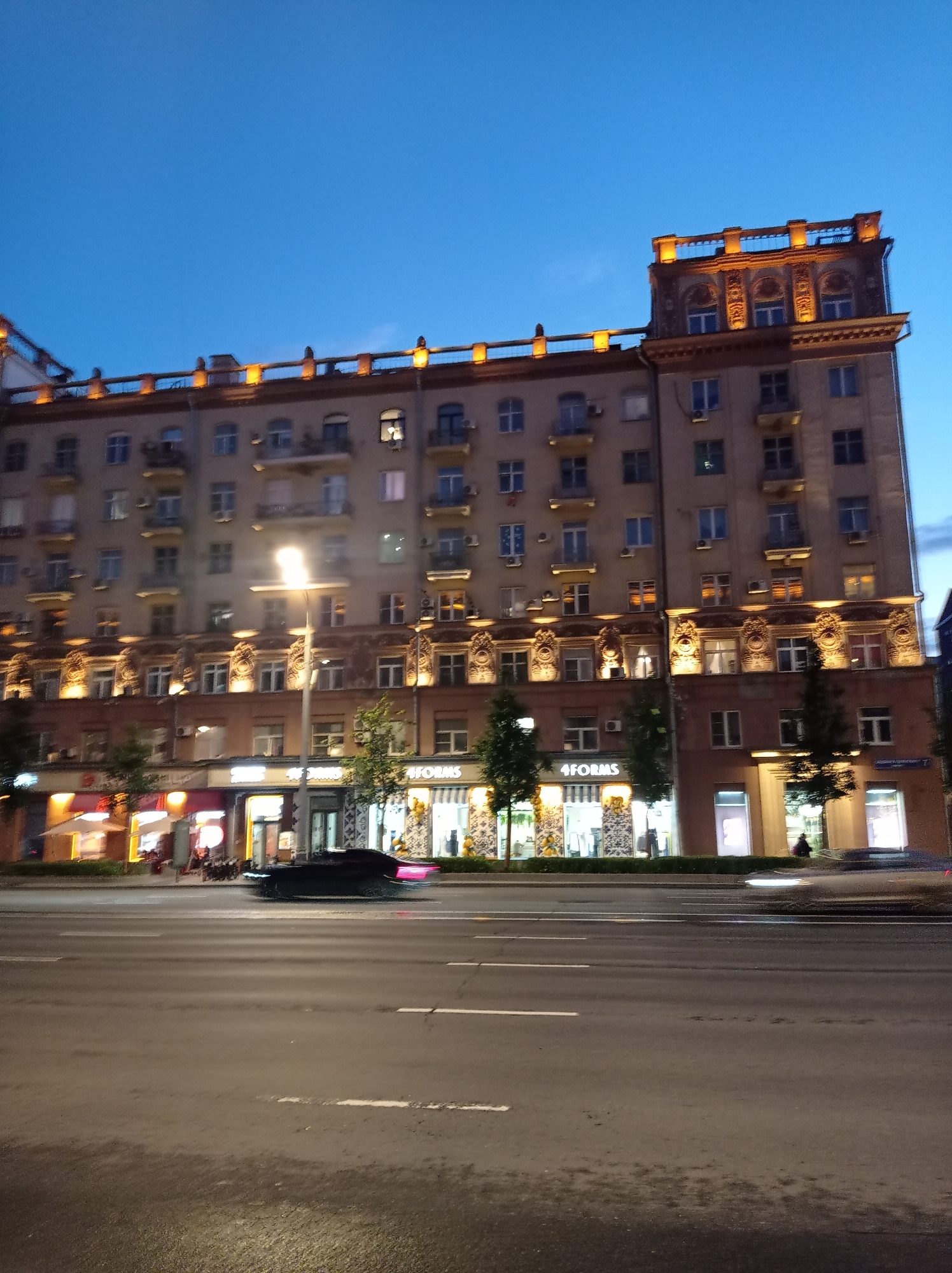 The image depicts a street scene in Moscow, Russia, at dusk or night. A multi-story building with numerous windows and ornate architectural details dominates the right side of the frame. The building is illuminated, with warm lights emanating from its upper floors and the storefronts at its base. Several businesses are visible on the ground level, including one with signage that reads "4FORMS". A street lamp stands in front of the building, casting a bright light. A dark-colored car is in motion on the road in front of the building, its taillights blurred due to movement. The road is paved with asphalt and marked with white lines. Trees are planted along the sidewalk in front of the businesses. The sky above is a deep blue, indicating twilight or early night. The overall impression is of an urban environment at night, with a focus on the illuminated building and the street activity.