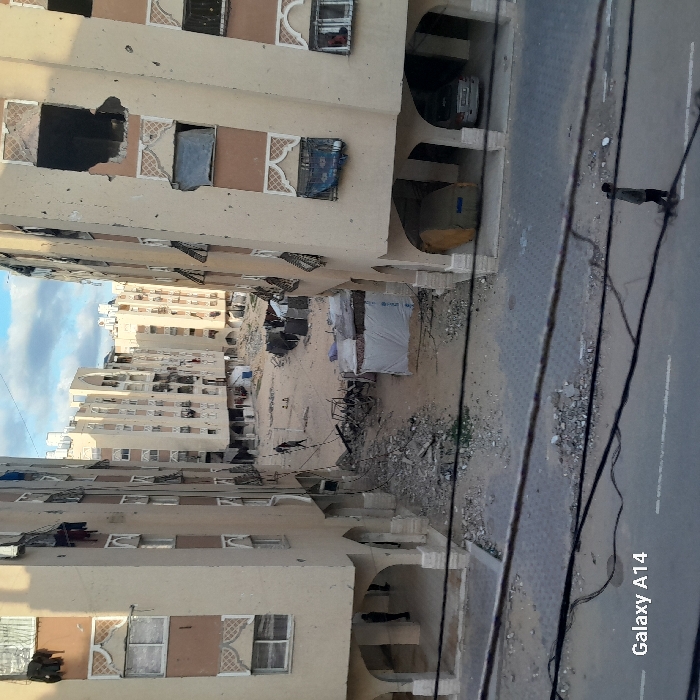 The image depicts a residential area in Khan Yunis, Palestinian Territory, characterized by severe structural damage. Two multi-story, beige apartment buildings frame a central, debris-strewn open space. Both buildings exhibit extensive damage, including numerous broken or missing windows, exposed interiors, and large holes in their facades. Debris, some resembling twisted metal and concrete, is scattered across the ground. In the central area, a small, makeshift shelter constructed from white tarp, bearing blue text, is visible amidst rubble. Further back, more similar apartment buildings, also showing signs of damage, line the street.
In the immediate foreground, a paved street runs horizontally, with scattered concrete fragments. Electrical wires cross the frame horizontally at varying heights. A lone individual, dressed in dark clothing, walks left-to-right on this street. In the damaged central area, two individuals in dark attire are seen walking away from the viewer. Under the arched ground floor of the left building, a dark figure stands. Under the arches of the right building, a silver sedan is parked alongside a large object covered in a yellow tarp. The sky above is partially cloudy.