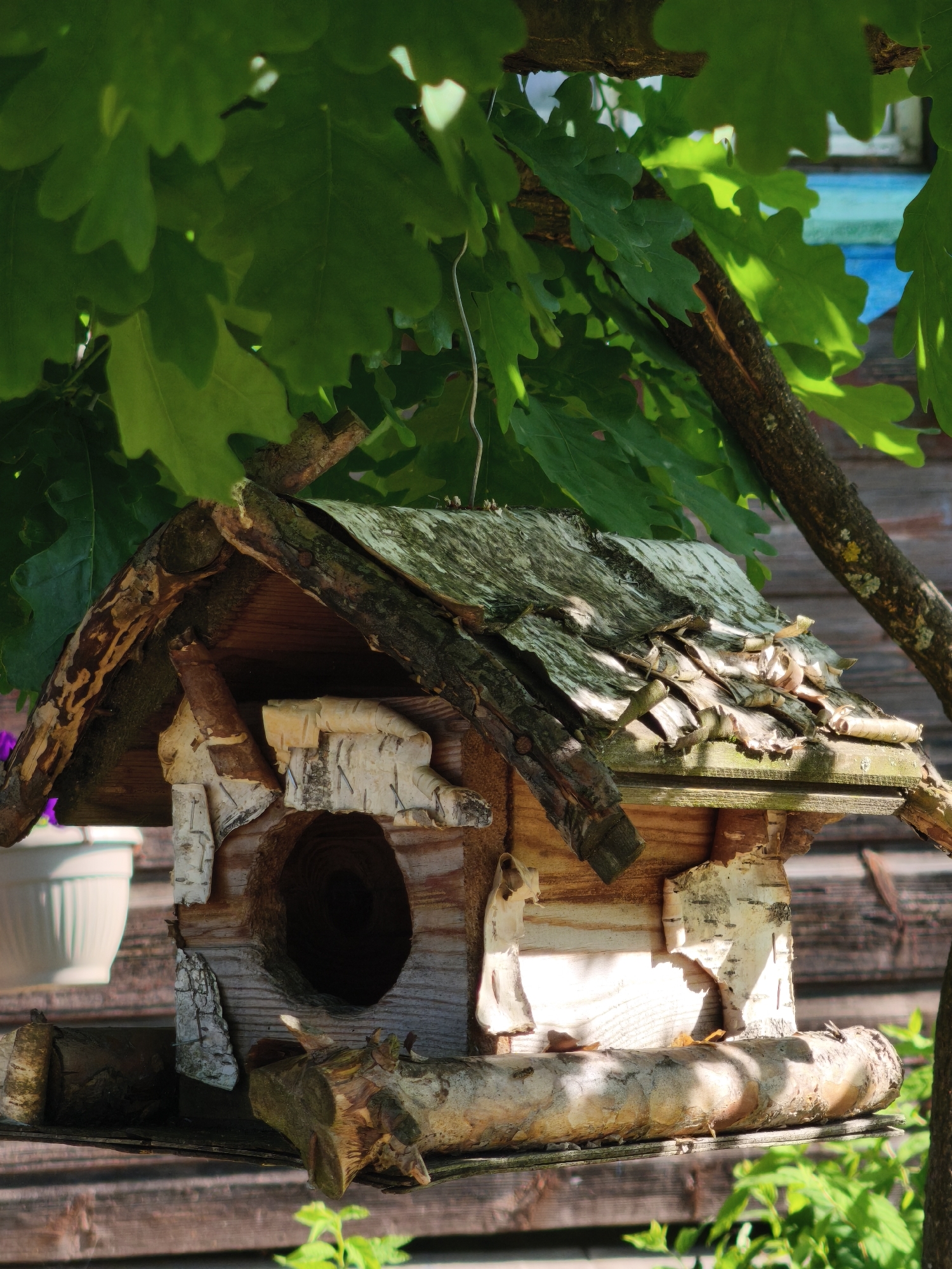 The image shows a rustic wooden birdhouse hanging from a tree branch, with lush green oak leaves overhead. The birdhouse is constructed from rough-hewn wood, bark, and birch bark, giving it a natural, organic appearance. It features a circular entrance hole and a perch made from a log. In the background, a weathered wooden fence and a glimpse of a blue structure are visible. The scene is bathed in dappled sunlight, creating a peaceful, outdoor setting. The location is identified as being in the city of Zhukovo, Russia.