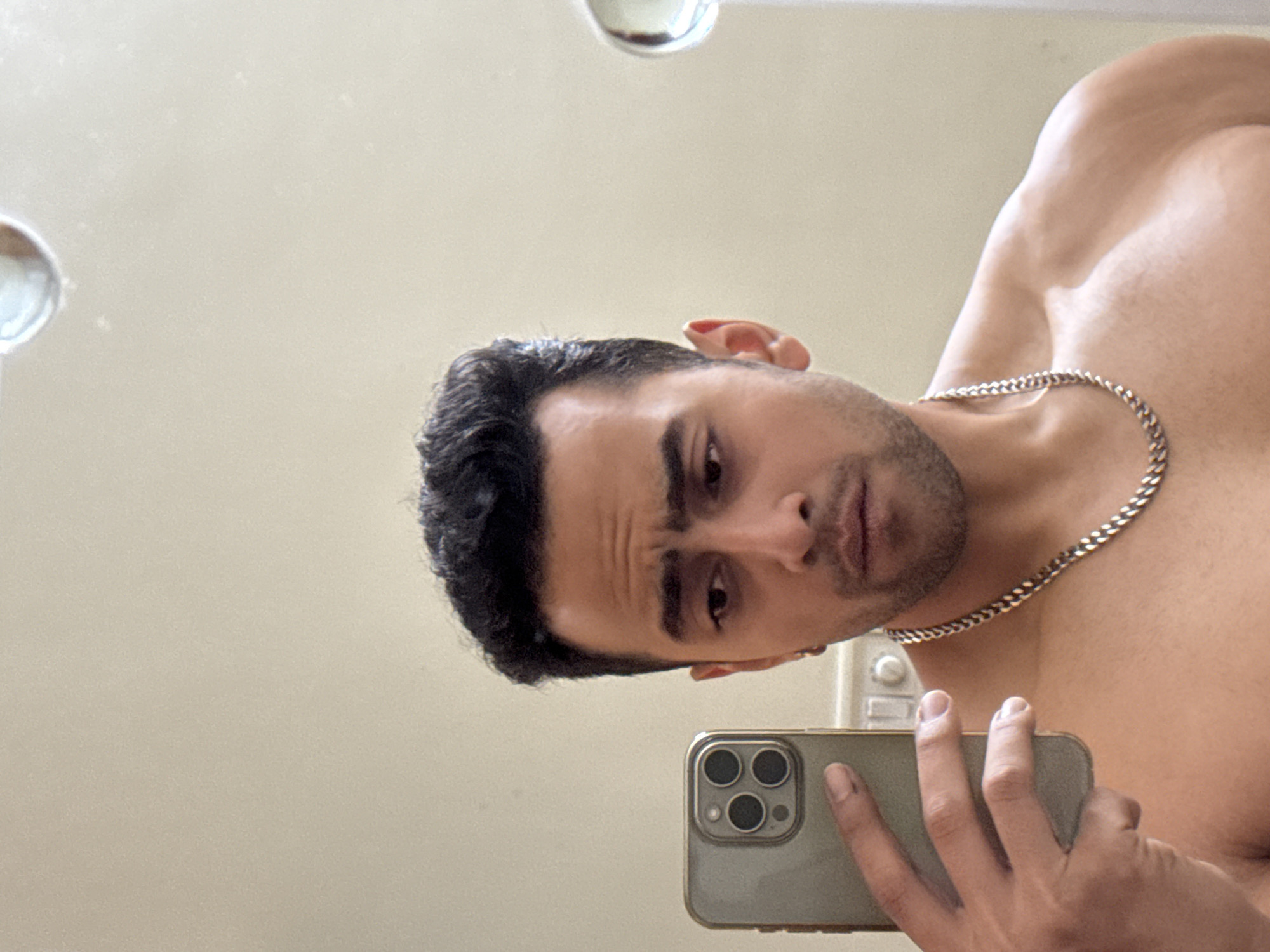 A person is seen in a mirror selfie, holding a smartphone with multiple camera lenses. The individual appears to be shirtless, wearing a chain around their neck. The background is a plain, light-colored wall with lighting fixtures, possibly in a bathroom setting. There is no indication of a location in Kolkata, India or any other identifiable location.