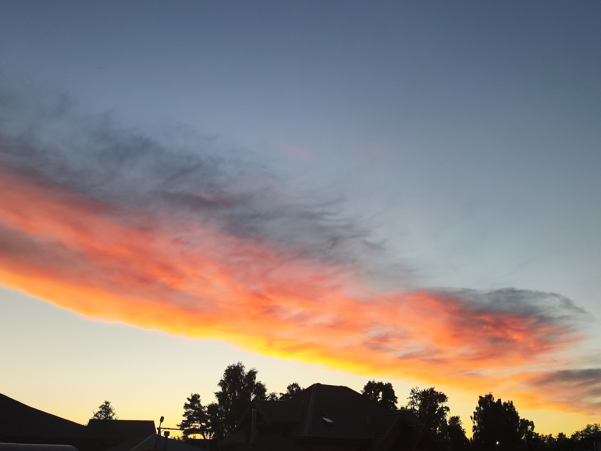 The image displays a vibrant sunset or sunrise over a residential area in Chaikovsky, Russia. The sky is the dominant feature, characterized by a stunning, elongated band of clouds stretching across the middle of the frame. This cloud formation is intensely illuminated in fiery shades of orange, red, and bright yellow by the low-lying sun. Above this colorful band, the sky transitions from a deep blue at the top to a lighter, paler blue, with some softer, darker, and wispier clouds scattered within the upper blue expanse. Below the main cloud band, the sky glows with a soft yellow light, indicating the sun's position near the horizon.
Silhouetted against the bright lower sky and the radiant clouds are the dark forms of several houses or buildings with sloped roofs, suggesting a typical suburban or rural architectural style. Numerous trees, both deciduous and coniferous, are also visible in dark silhouette along the horizon line, some with distinct branches and foliage, others appearing as dense masses. A single street light or similar pole structure can be faintly discerned in silhouette on the right side, between the trees and houses. The overall scene conveys a sense of dramatic natural beauty and tranquility at dusk or dawn. No text is visible in the image.