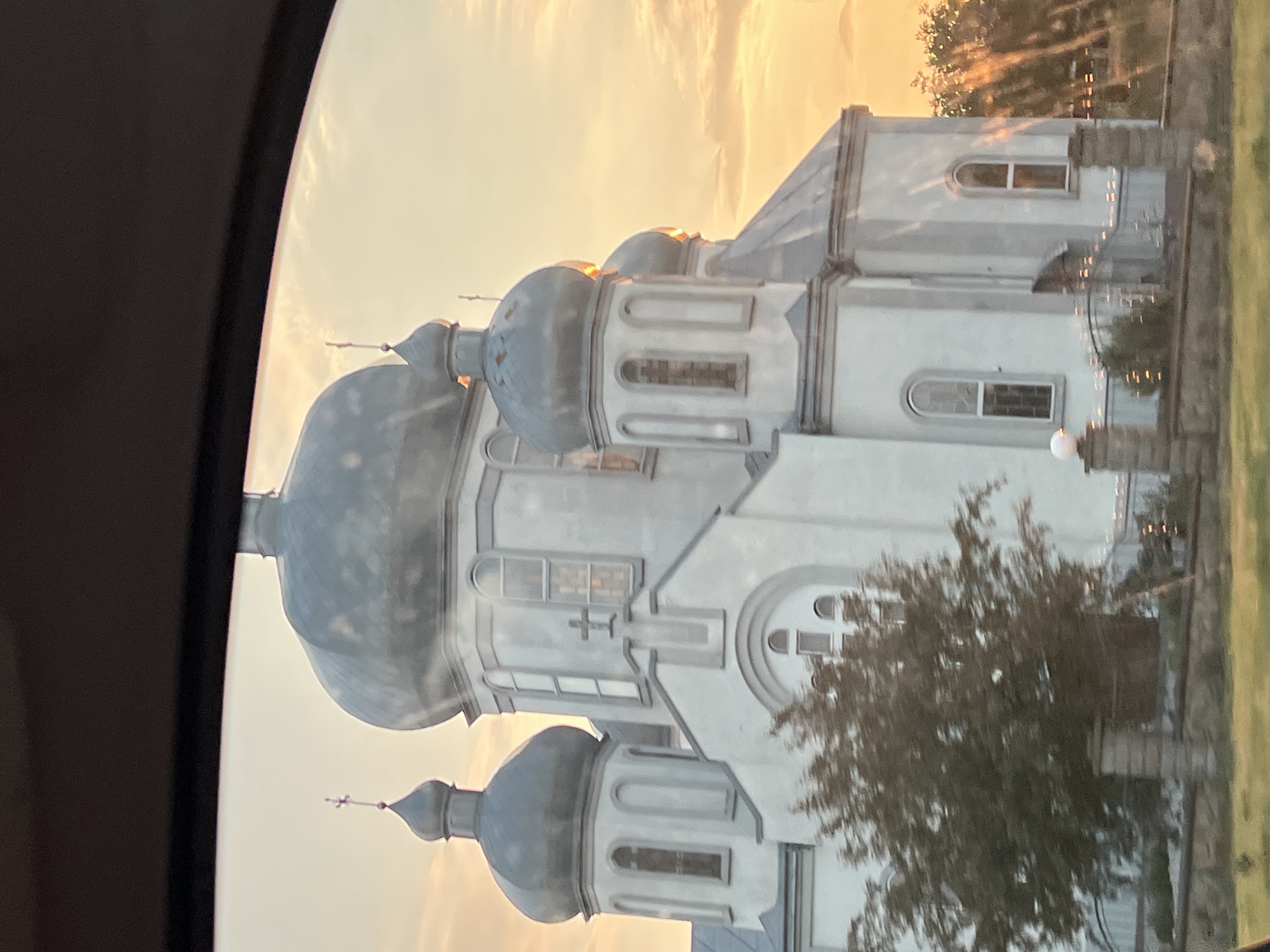 The media file presents a striking view of a large, multi-domed church or cathedral, likely an Eastern Orthodox edifice, set against a vibrant golden sky. The building's exterior is light-colored, appearing white or pale grey, featuring numerous dark grey, onion-shaped domes. Several domes reflect the warm, golden light of either sunrise or sunset, with golden highlights prominent on their rounded surfaces and the slender spires, some of which are topped with crosses.
The sky is a clear, warm expanse of yellow and orange hues, characteristic of the 'golden hour'. In the foreground and midground, green trees with dense foliage partially obscure parts of the church, and a strip of green grass is visible along the right edge of the frame. The image appears to have been captured from inside a vehicle, as suggested by the dark, curved frame on the left, resembling a car window.
The scene is an exterior shot of the religious building, located in Bila, Ukraine. No people or discernible text are visible, emphasizing the architectural grandeur and the serene natural light of the setting.