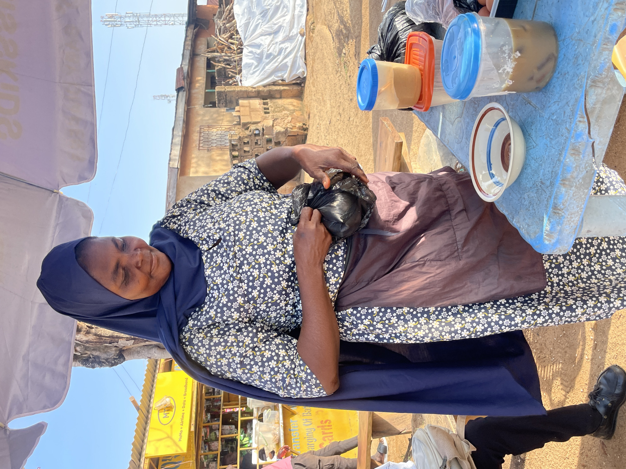 A woman wearing a dark blue hijab and a floral patterned dress stands under a white canopy, looking down at a black plastic bag she is holding. She wears a brown apron over her dress. A blue plastic container filled with a light brown liquid and ice is on a blue table in front of her. Next to it is a small bowl with red and blue stripes. Behind the table, on a shelf, are more plastic containers and black bags. In the background, a yellow sign with the logo "MTN" is visible, along with some buildings and cell towers. The scene appears to be an outdoor market stall in Jos, Nigeria.