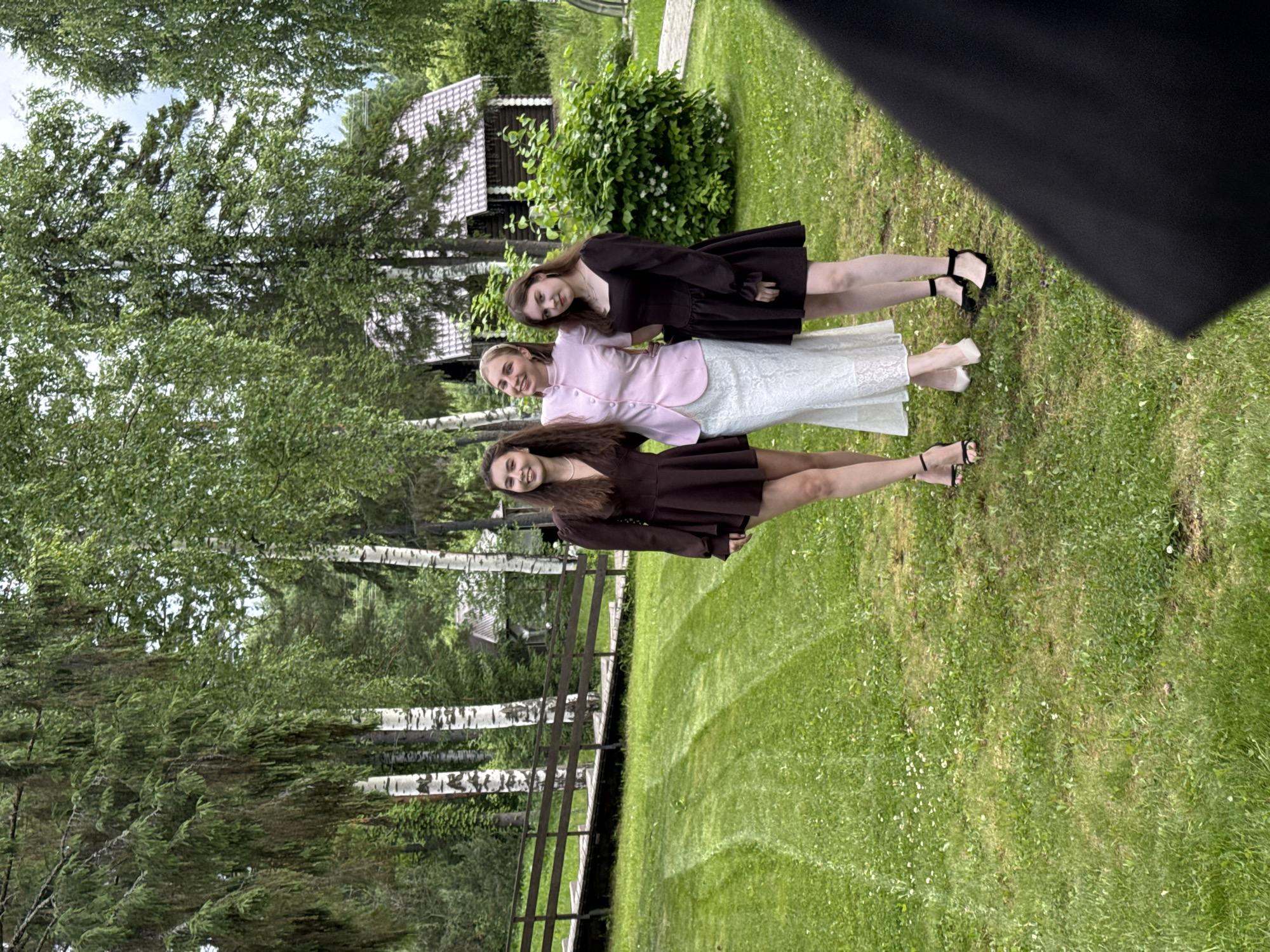 In Votkinsk, Russia, three young women are posing for a photo outdoors on a bright, overcast day, likely in the early afternoon. They are standing on a grassy lawn in front of a wooded area with birch trees. Behind them, a rustic wooden building with a white, patterned roof and decorative trim is visible. The women are dressed formally, suggesting a special occasion like a graduation or a party.
The woman in the middle wears a light pink jacket over a white lace skirt, while the other two are in matching dark brown dresses. All three are wearing heeled sandals. The woman on the left is looking directly at the camera with a slight smile, while the other two are looking slightly off-camera, also with smiles. The lighting is diffused, indicating it's not direct sunlight, and the overall atmosphere is relaxed and cheerful. The setting appears to be a rural or semi-rural environment, with lush greenery and a sense of privacy. There is no visible text in the image.