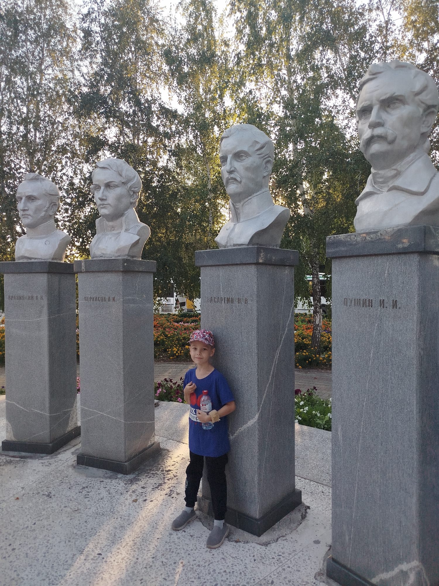 The image depicts a group of four busts on stone pedestals, with a young boy standing in front of the third pedestal from the left. The busts appear to be of historical figures, possibly writers or poets, given the inscriptions on the pedestals which are in Cyrillic. The bust on the third pedestal has the inscription "ПУШКИН И. И." which translates to "Pushkin I. I.". The busts and pedestals are made of stone, and the surrounding area is paved with stone tiles. There are trees in the background with dappled sunlight filtering through them. The boy is wearing a blue t-shirt, black pants, a baseball cap, and grey shoes. He is holding a bottle of water and a small toy. The scene is likely outdoors, possibly in a park or public square in the city of Yalutorovsk, Russia, given the presence of the bust of the renowned Russian poet Alexander Pushkin, who has significant ties to the city. The overall atmosphere is serene, with the golden hour sunlight casting long shadows.