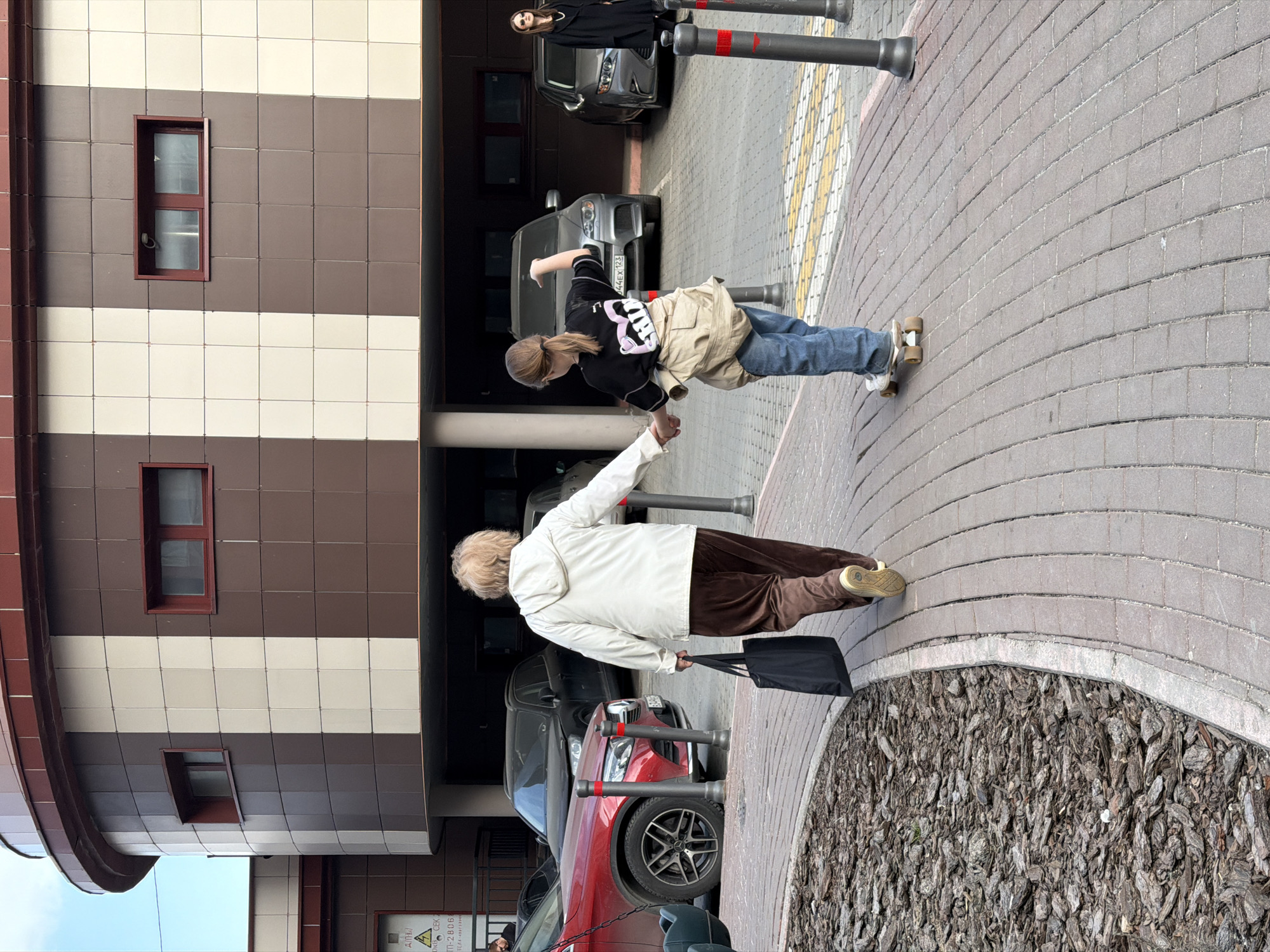 In Moscow, Russia, an elderly woman in a white jacket and brown pants walks along a paved sidewalk, carrying a black bag. A younger woman in a black shirt, blue jeans, and a tan jacket reaches out to assist her. They are near a building with a brown and white striped facade and windows with maroon frames. Several cars are parked nearby, including a red SUV and a gray sedan. Bollards line the sidewalk.