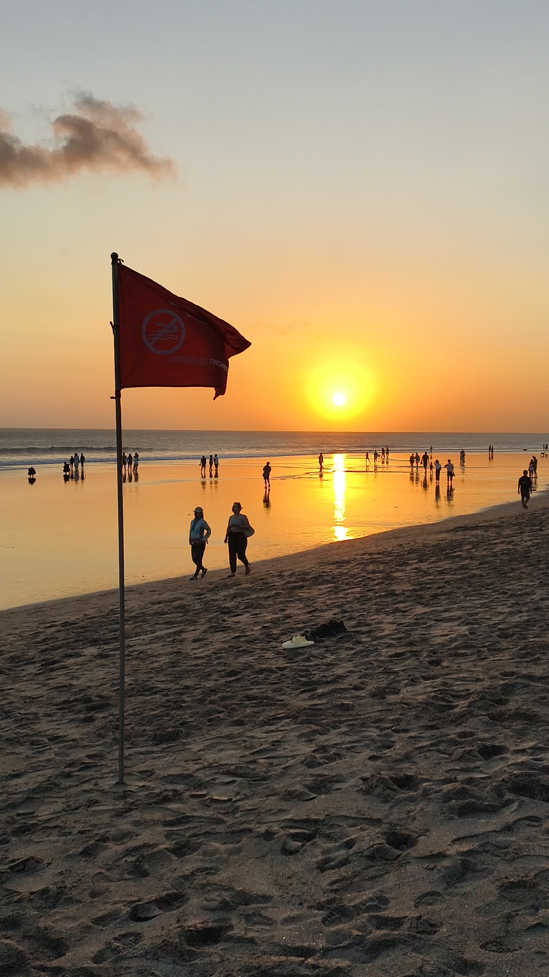 The image depicts a beach scene at sunset in North Kuta, Indonesia. The sky is a gradient of soft yellow and orange, with the sun a bright orb setting on the horizon. The calm ocean reflects the warm hues of the sunset, creating a shimmering effect on the wet sand. Numerous people are scattered across the beach and in the shallow water, engaged in various activities such as walking, standing, and observing the sunset. In the foreground, a red flag with a white symbol indicating "swimming prohibited" flies from a pole on the sandy beach. Two individuals are walking on the sand in the foreground, with their backs partially turned to the camera. The sand itself is a dark, textured surface with footprints visible. A pair of white and black shoes are placed on the sand in the mid-ground. The overall atmosphere is peaceful and serene, capturing the end of a day at the beach.
