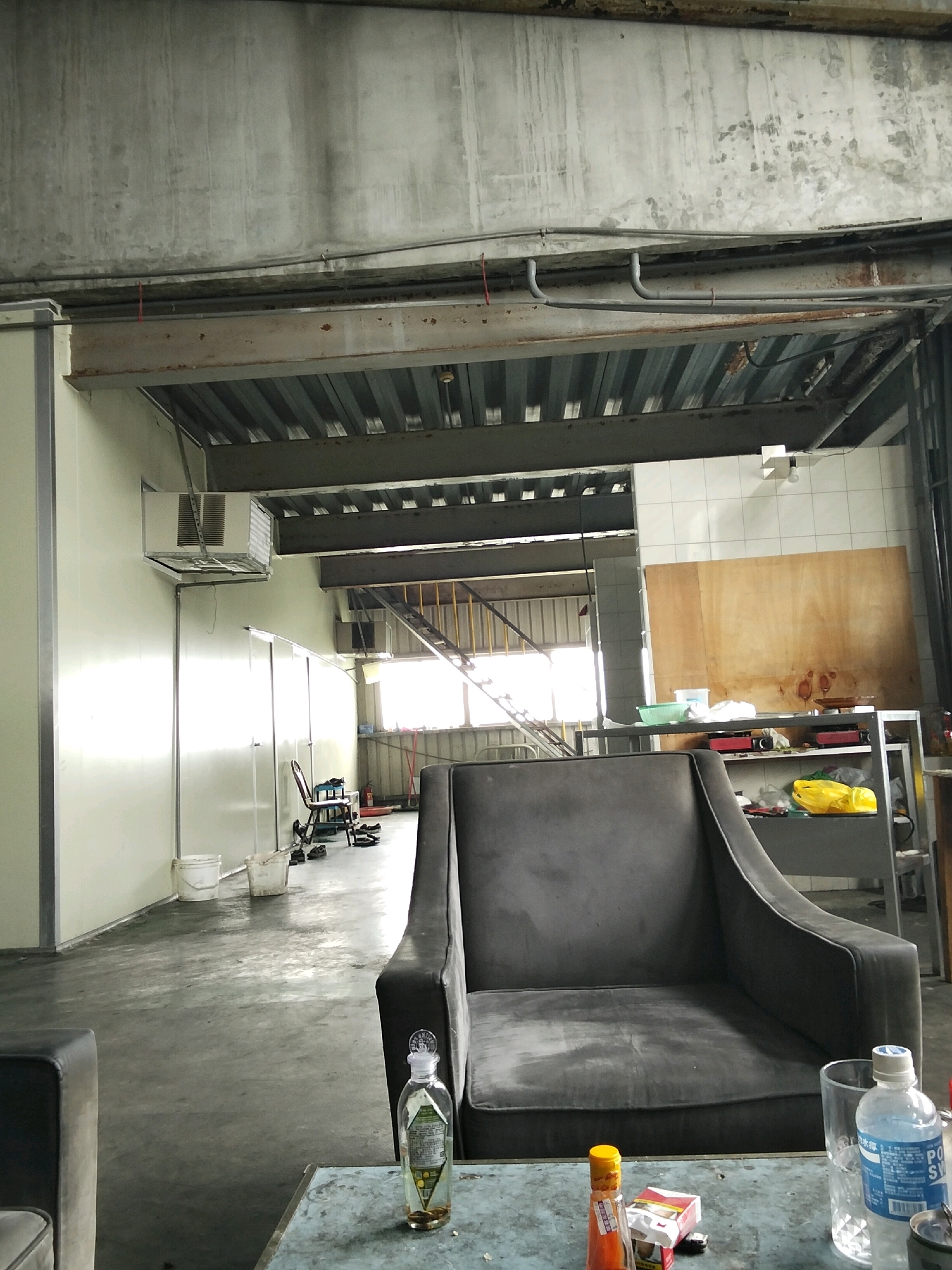 The image depicts an indoor scene, possibly a storage or utility area, with a concrete floor and exposed ceiling beams. The environment appears somewhat unkempt. In the foreground, there is a dark gray armchair and a low table with several objects on it, including a bottle of liquid with coins inside, a bottle of what appears to be hot sauce, a pack of cigarettes, and a glass. To the left of the armchair, another dark gray seating area is partially visible. In the background, there are white walls, a metal staircase leading upwards, and large windows that allow natural light to enter. Various items are scattered around, such as buckets, a chair, and shelving units containing what might be tools or supplies. The overall impression is of a functional, yet cluttered, space. No people are visible in the image.