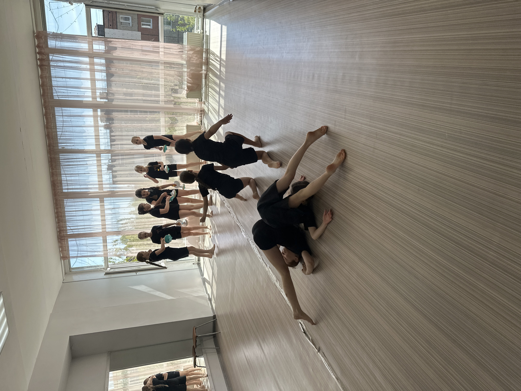 The image shows a dance studio in Votkinsk, Russia. A group of about ten people, mostly women, are in the room. They are dressed in black, some holding small blue objects. They appear to be engaged in a dance or exercise class. Some are standing near a wall with large windows, while others are on the floor, performing various poses. The room has a light-colored, wood-grain floor, and there's a ballet barre visible in the lower left corner of the image. The windows have light-colored curtains and offer a view of a building outside.