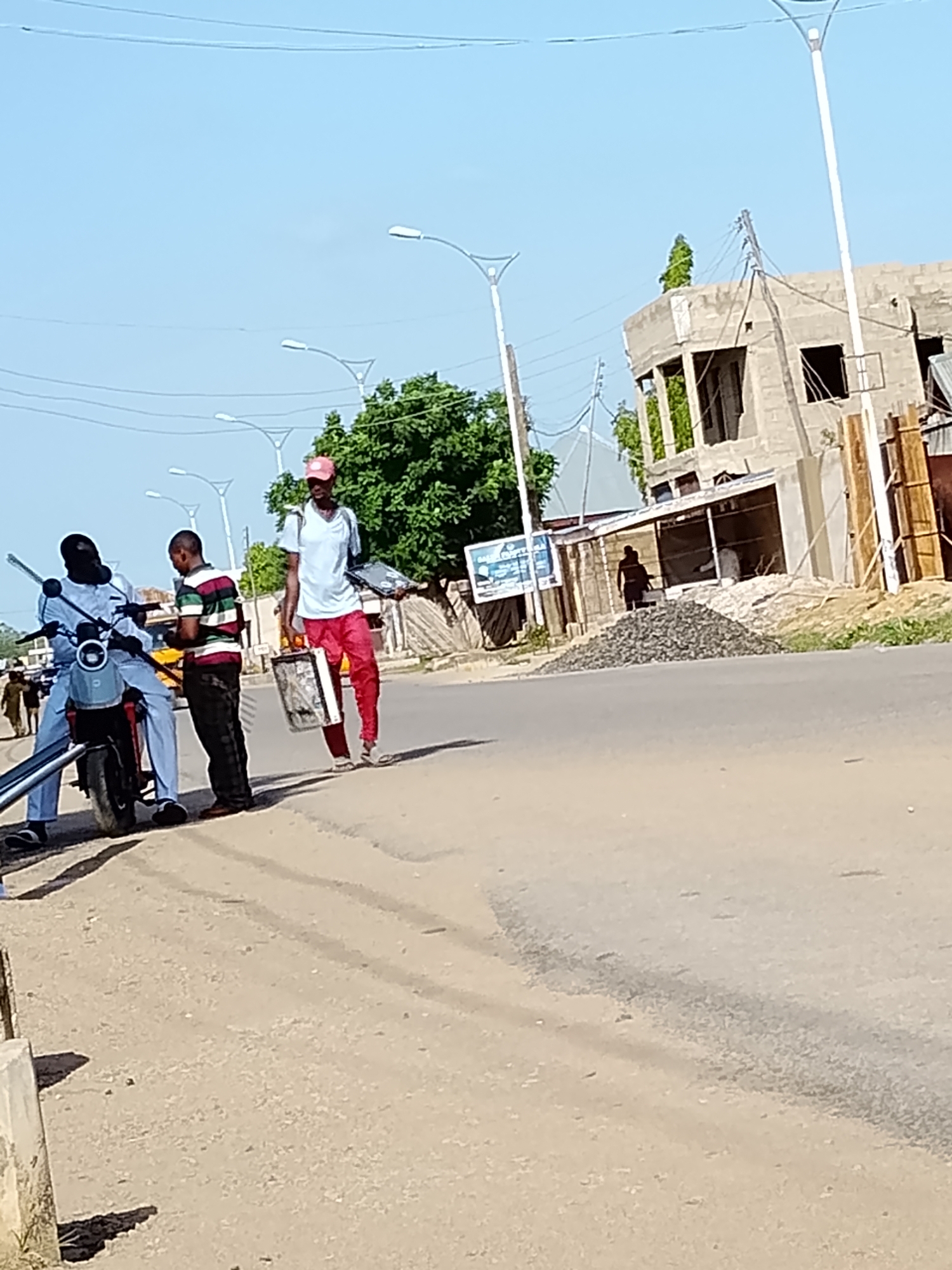 In Wauro Leggal, Nigeria, on a bright, sunny day, three men are observed near a street. The man on the left is seated on a light blue electric scooter, wearing a light blue tunic and matching trousers. To his right stands a man in a horizontally striped green, white, and red shirt and dark trousers, holding a small yellow object. The third man, standing further to the right, wears a red baseball cap, a light blue t-shirt, and bright red trousers. He carries a rectangular object, possibly a laptop, and a woven basket. Behind them, a building under construction is visible, with scaffolding and piles of gravel. A few trees and utility poles are also present in the background. The scene is captured on a paved road, with shadows indicating the strong sunlight.