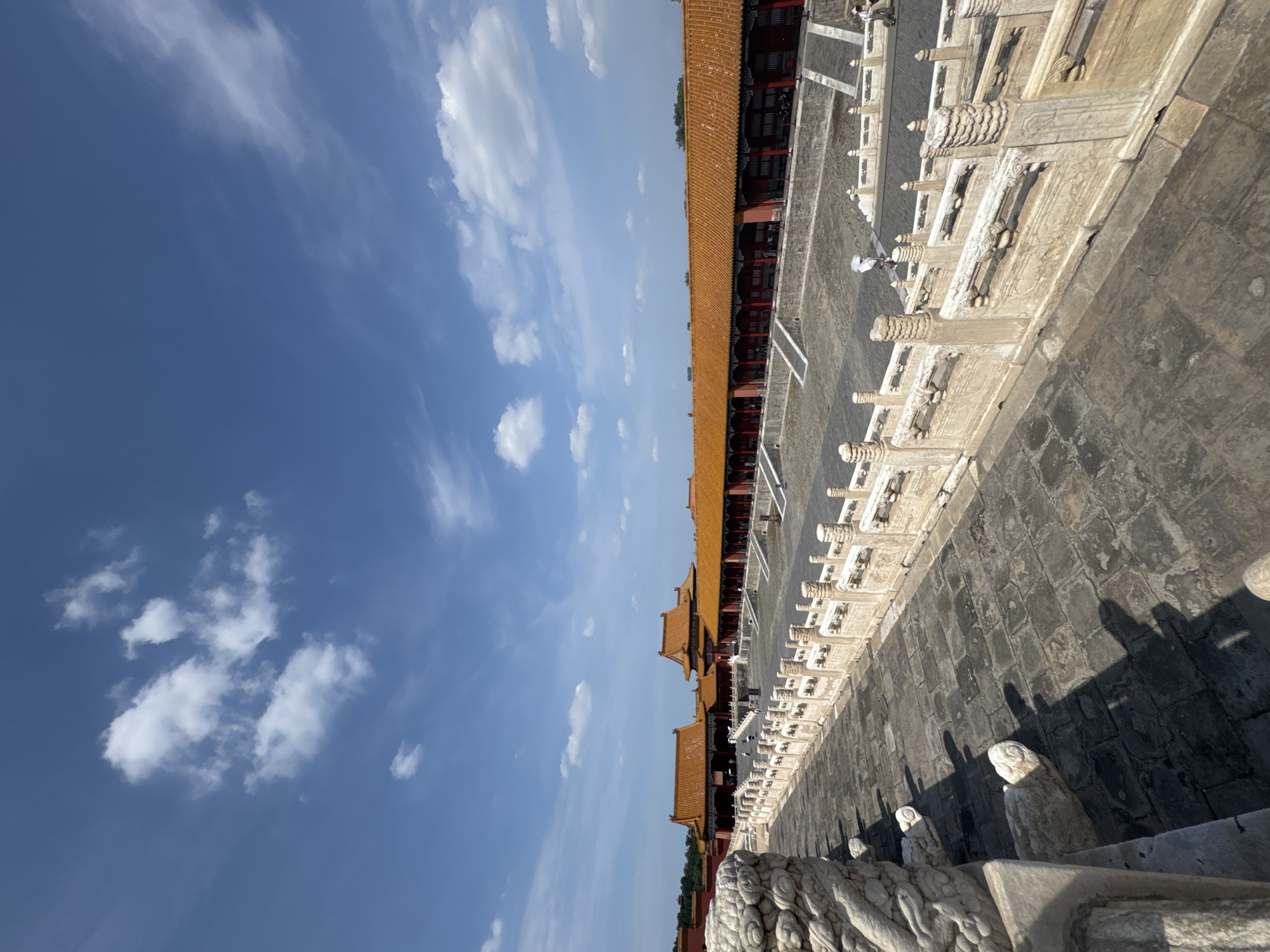 This image depicts a wide courtyard within the Forbidden City, located in Dongcheng District, China. The foreground is dominated by a stone pathway and an ornate white marble balustrade with intricately carved decorative elements, including what appear to be dragon motifs. The pathway is paved with large, dark gray stone slabs. Beyond the balustrade lies a vast, open courtyard.
In the mid-ground, a long, red building with a traditional orange-tiled roof stretches horizontally across the frame. This building features numerous red columns supporting a covered walkway. Several people are visible within the courtyard, some walking, and one person wearing a white hat appears to be on a bicycle. The architecture is characteristic of traditional Chinese imperial palaces.
The background shows more buildings with similar orange roofs and darker walls, suggesting a complex of structures. The sky above is a clear blue with scattered white cumulus clouds. The overall scene conveys a sense of historical grandeur and open space within a significant cultural landmark.