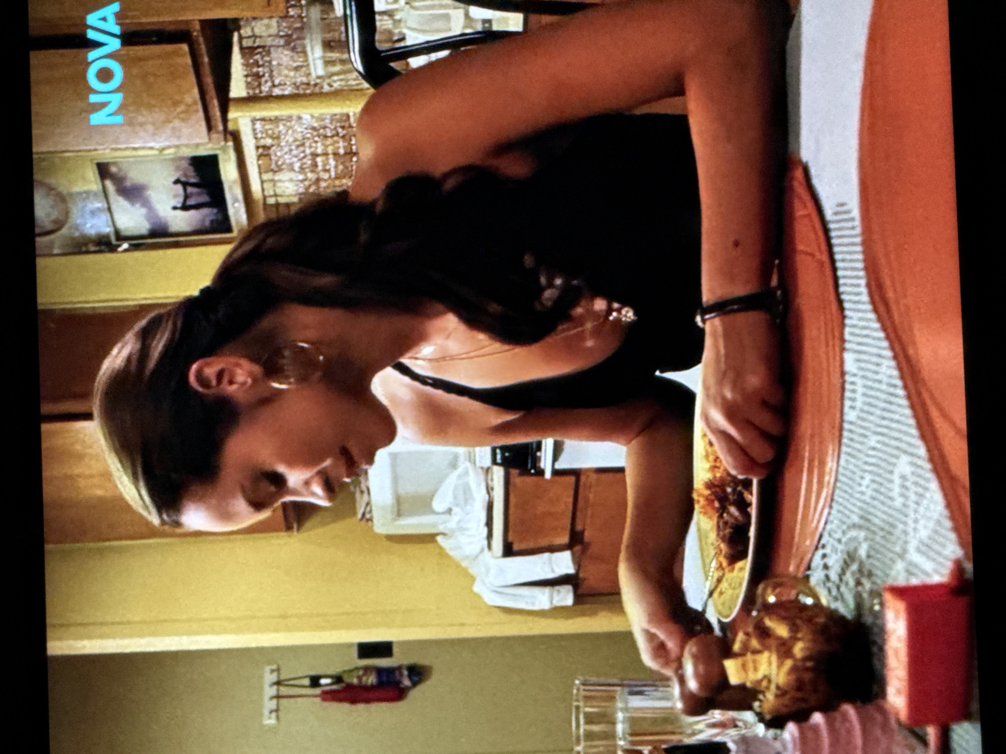 The image shows a woman sitting at a table in what appears to be a kitchen. She is wearing a black tank top and large hoop earrings. Her hair is pulled back in a ponytail. She is reaching for a salt and pepper grinder on the table, which is set with plates of food. There is a glass of water on the table as well. In the background, there are kitchen cabinets, a microwave, and a framed picture on the wall. The logo "NOVA" is visible in the upper right corner of the image. The scene does not provide any information about the city of Delmenhorst, Germany.