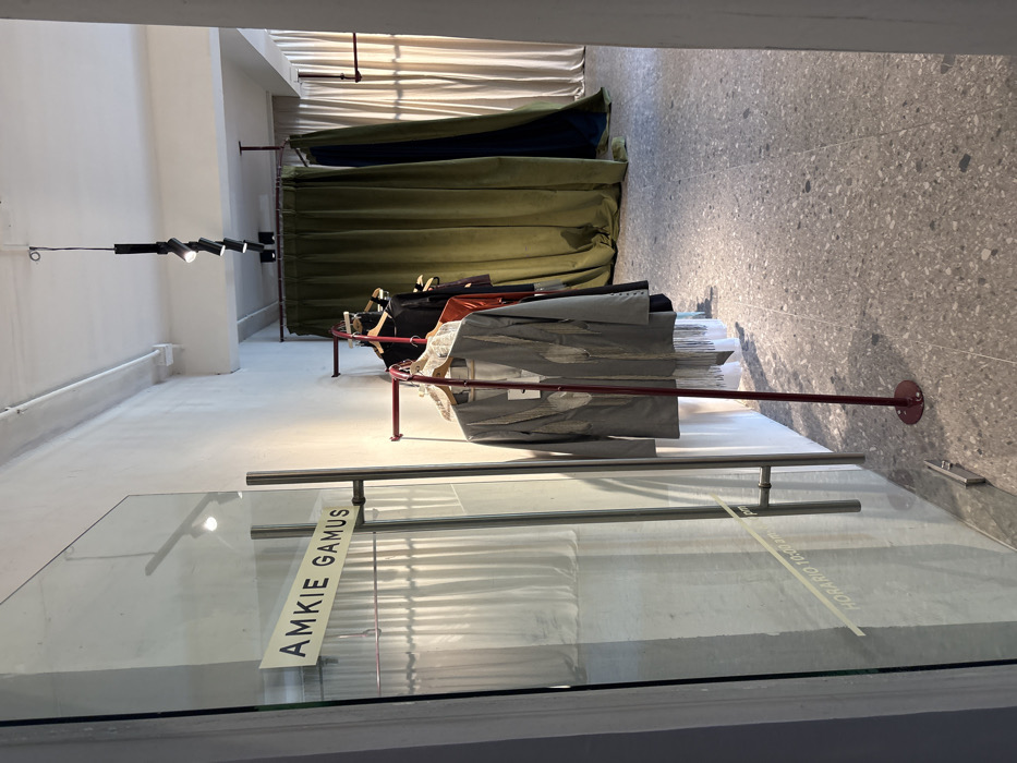A store window showcases clothes on a red, curved rack. Gray blazers are at the front, followed by darker garments. Behind the rack, curtains of olive green and beige provide a backdrop. A glass panel displays the name "AMKIE GAMUS" vertically. The floor transitions from white near the window to a speckled gray further inside the store. A modern lighting fixture with multiple cylindrical lights is attached to the ceiling.