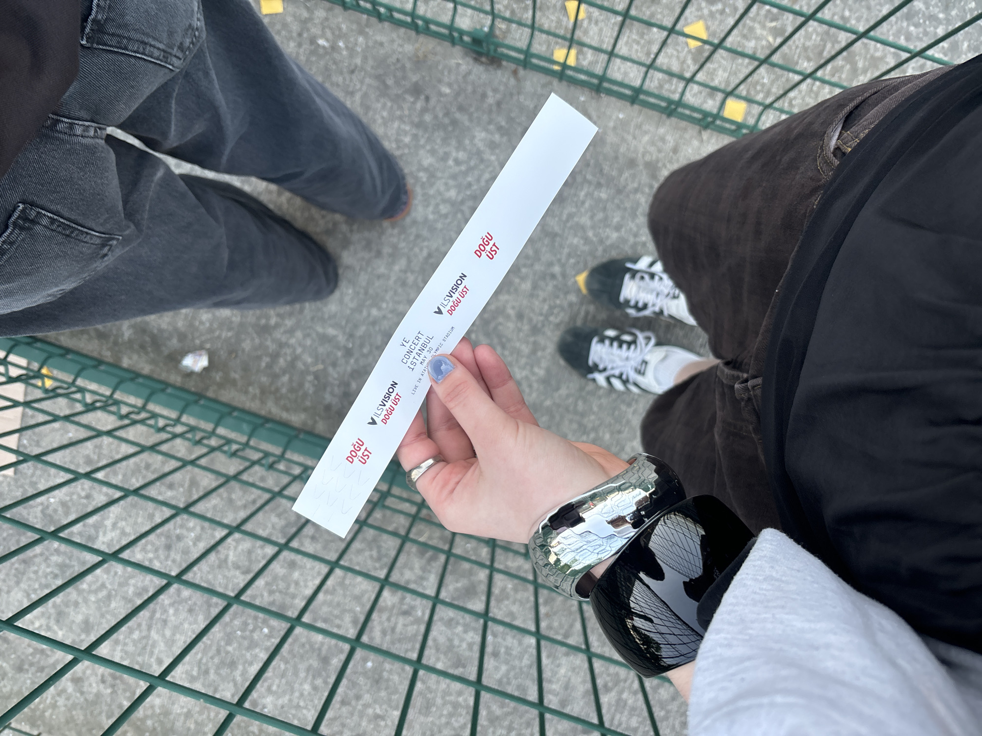 An overhead shot shows two individuals standing on a paved surface through which a green wire grid is visible. One individual is wearing gray jeans, while the other is wearing brown pants. The person closer to the camera is holding a concert ticket in their hand, adorned with two bracelets, one silver and one black. The ticket bears the inscription "YE CONCERT ISTANBUL MAY 30 LIVE IN ATATURK OLYMPIC STADIUM," with "VILSVISION DOGU UST" also printed on it. The location is potentially in Başakşehir, Turkey, as Ataturk Olympic Stadium is in this area.