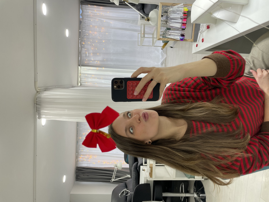 A woman poses for a selfie, tilting her head to the side while pouting. She wears a red and brown striped sweater and a large red bow in her hair. She holds up a black phone with a red pop socket. The photo appears to be taken in a nail salon, with manicure tools and salon chairs visible in the background. White curtains decorated with fairy lights hang in the background.