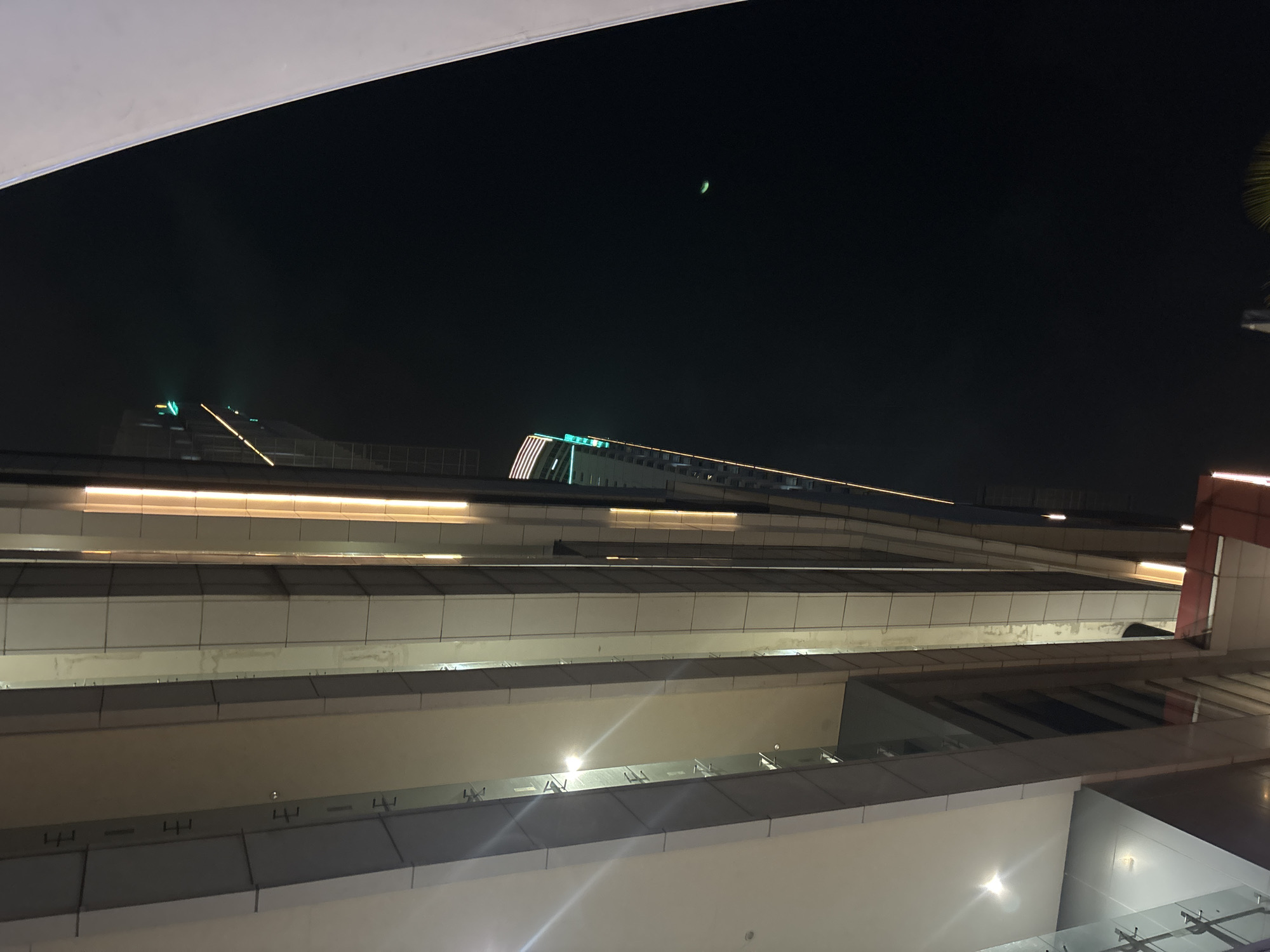 The image, taken at night in North Jakarta, Indonesia, showcases a portion of a modern high-rise building with a striking facade. The building is illuminated by vertical strips of warm, orange light running along its exterior, contrasting with the dark night sky. Some sections of the building feature a sleek, metallic finish, while others are clad in light-colored panels. On the far right, a large, white, curved structure partially obscures the view, suggesting a pedestrian walkway or a section of an adjacent building. In the distance, the upper floors of another skyscraper are visible, adorned with bright green lights. A faint, crescent-shaped green object, possibly a distant light or a celestial body, is discernible in the dark expanse of the sky. The perspective is looking upwards, emphasizing the height and architectural design of the buildings. No people are visible in the frame.