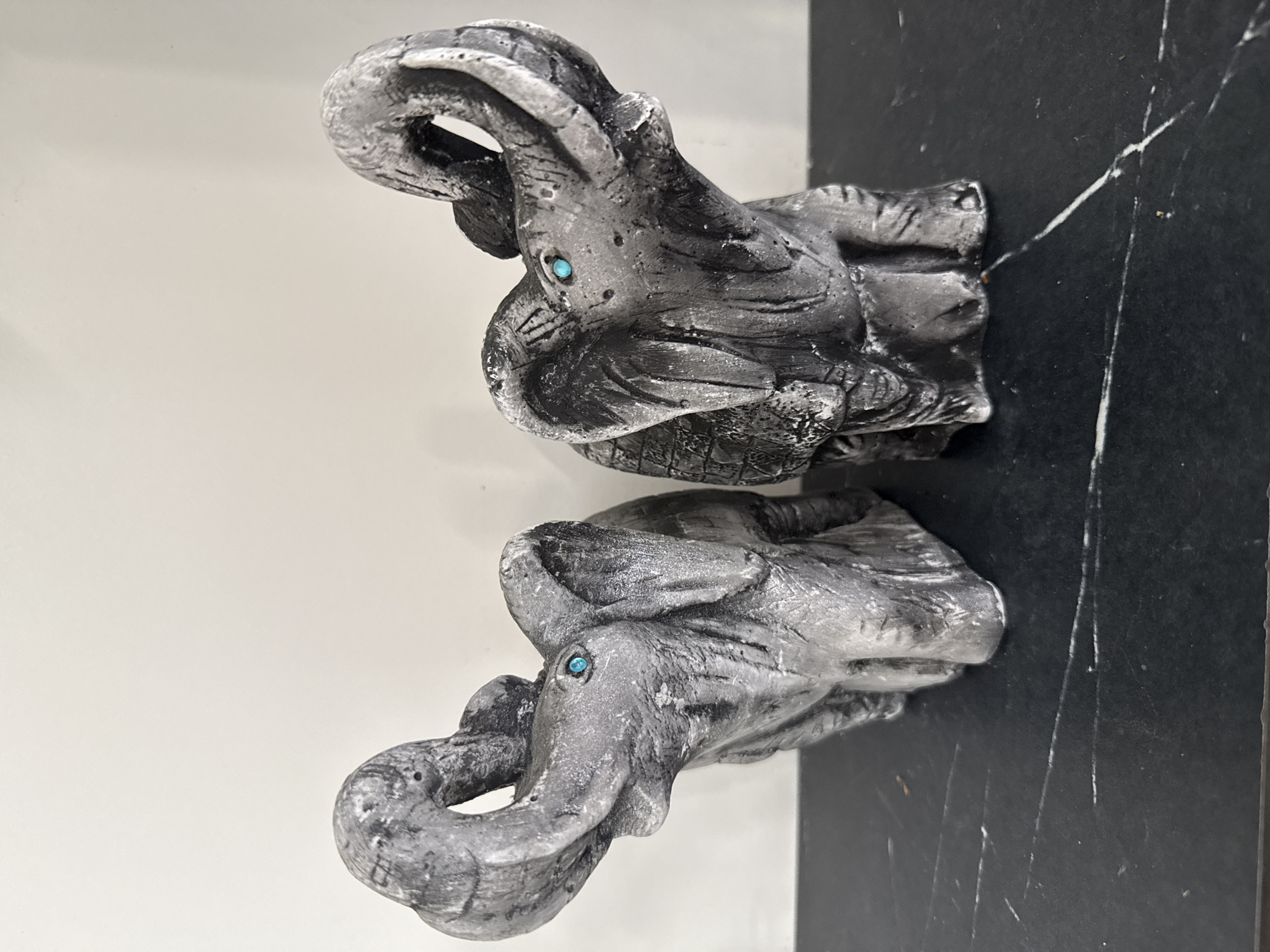 Two grey elephant figurines with upturned trunks and small turquoise eyes are positioned side-by-side on a dark, reflective surface, possibly a countertop or shelf. The figurines appear to be made of stone or a similar textured material, with visible lines and imperfections suggesting a handcrafted quality. The background is a plain, light-colored wall. The scene is indoors. The provided image does not contain information to determine its location in Khokhryaki, Russia.