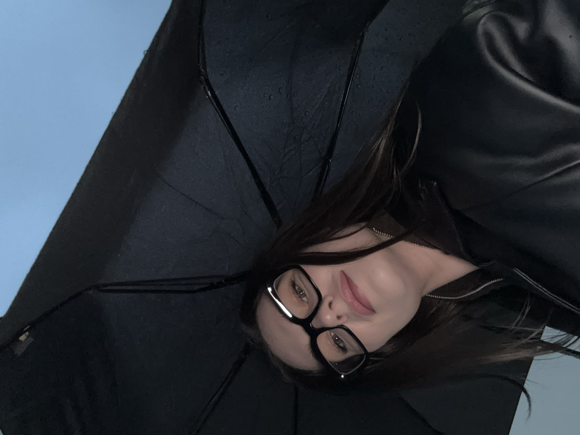 A woman with dark hair and glasses is holding a black umbrella, possibly in Chaikovsky, Russia, although location cannot be definitively determined from the image. She is wearing a black jacket with a visible zipper. The background is a light blue, suggesting a sky. The umbrella has water droplets on it, indicating rain.