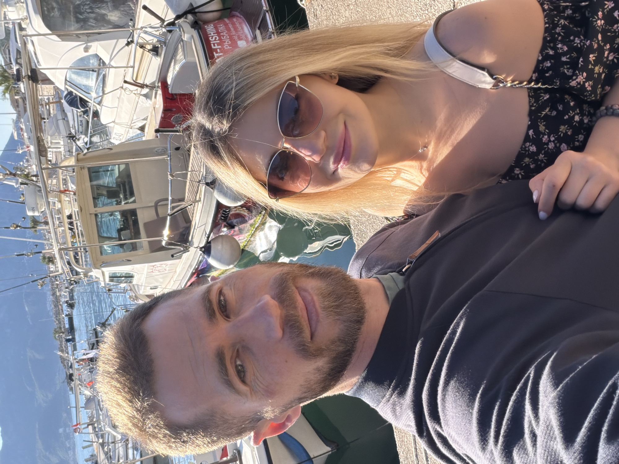 A selfie captures a man and a woman against the backdrop of a marina, presumably in Budva, Montenegro, given the appearance of the architecture and boats. The man, positioned at the bottom of the frame, has light brown hair, a beard, and is wearing a dark-colored long-sleeved shirt. He looks directly at the camera with a slight smile. Above him, the woman has blonde hair, sunglasses, and is wearing a floral dress with a white strap. She is also smiling at the camera. Behind them, multiple boats are docked in a harbor. The background indicates a bright sunny day, with reflections visible in the water.