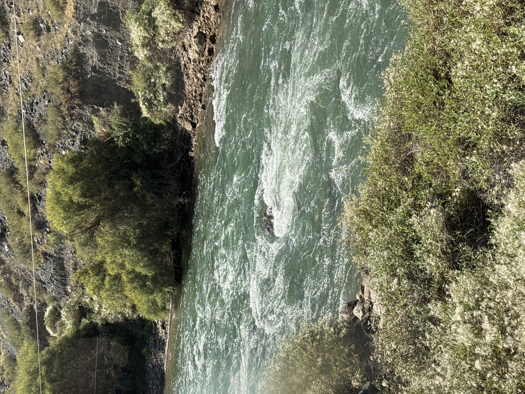 The image depicts a fast-flowing, greenish river in a mountainous, natural environment. The riverbanks are rocky and covered with dense vegetation, including trees and bushes with fluffy white flowers. A dark rock is visible in the middle of the turbulent water, causing a significant disturbance. In the background, a steep, rocky hillside is sparsely covered with vegetation. A thin, light-colored wire stretches horizontally across the upper part of the scene, possibly part of a power line or fence. The overall impression is a rugged, natural landscape with a powerful river. The location is identified as Unknown, Kyrgyzstan.