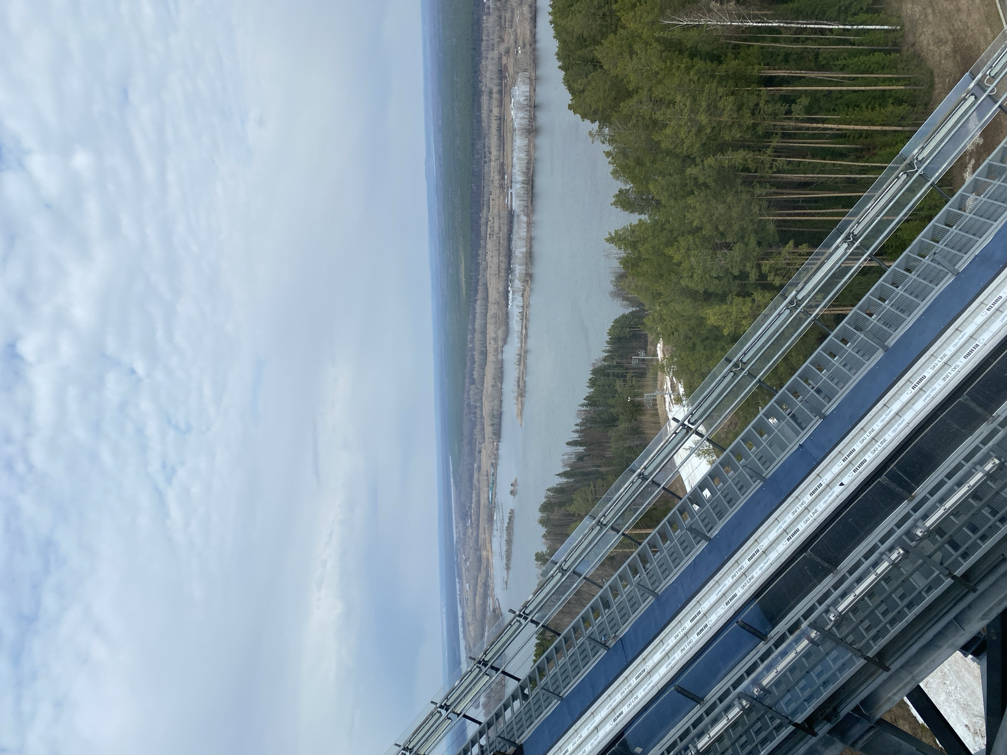 Here's a detailed description of the image:
**General Scene:** The image presents an aerial view of a landscape. The vantage point appears to be from a high structure, possibly a tower or elevated platform.
**Landscape:** A river or large body of water stretches through the scene. On both sides of the river, there are patches of forest. The trees seem to be a mix of deciduous and evergreen types.
**Structure:** Dominating the foreground and middle ground is the metallic structure from which the photograph seems to be taken. It consists of a network of metal supports, grids, and possibly rails. Text is visible on parts of the structure "REHAU SKI-LINE" suggesting it is for a winter sports activity, likely for skiing.
**Weather:** The sky is overcast with clouds.
**Location:** This image shows the location in the city of Прикамский, Russia.