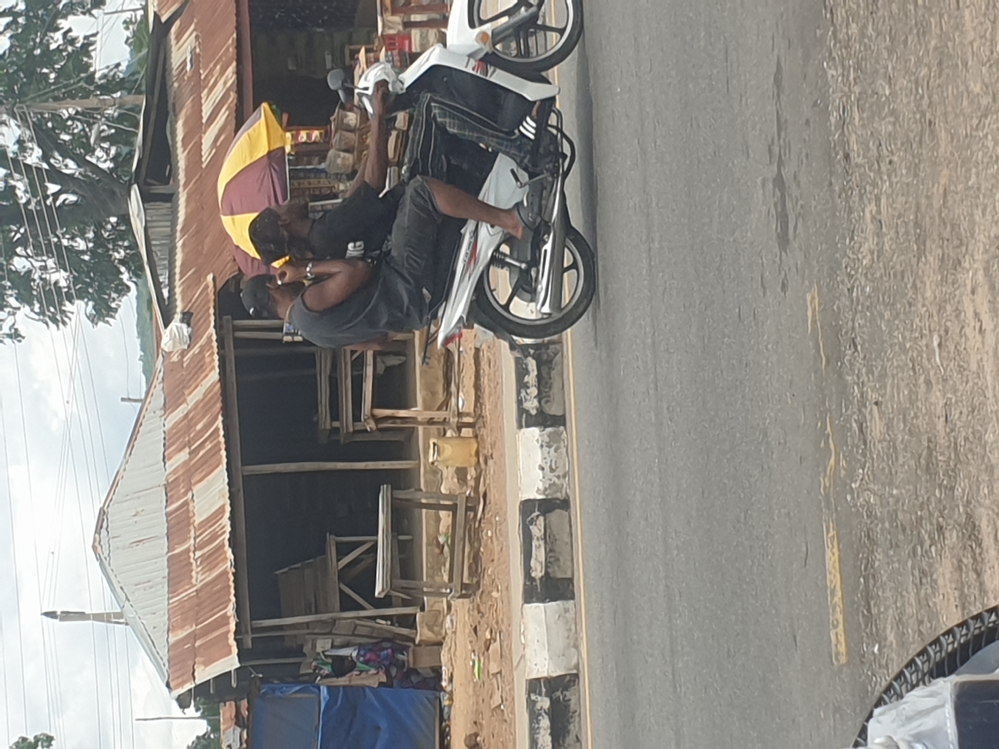 Two men are sitting on a white motorcycle parked on the side of a road. The man in front is wearing a gray tank top and a black baseball cap. The man behind him is wearing a black shirt. They are in front of a building with a corrugated metal roof and a yellow and burgundy umbrella. The location is Unknown, Unknown.