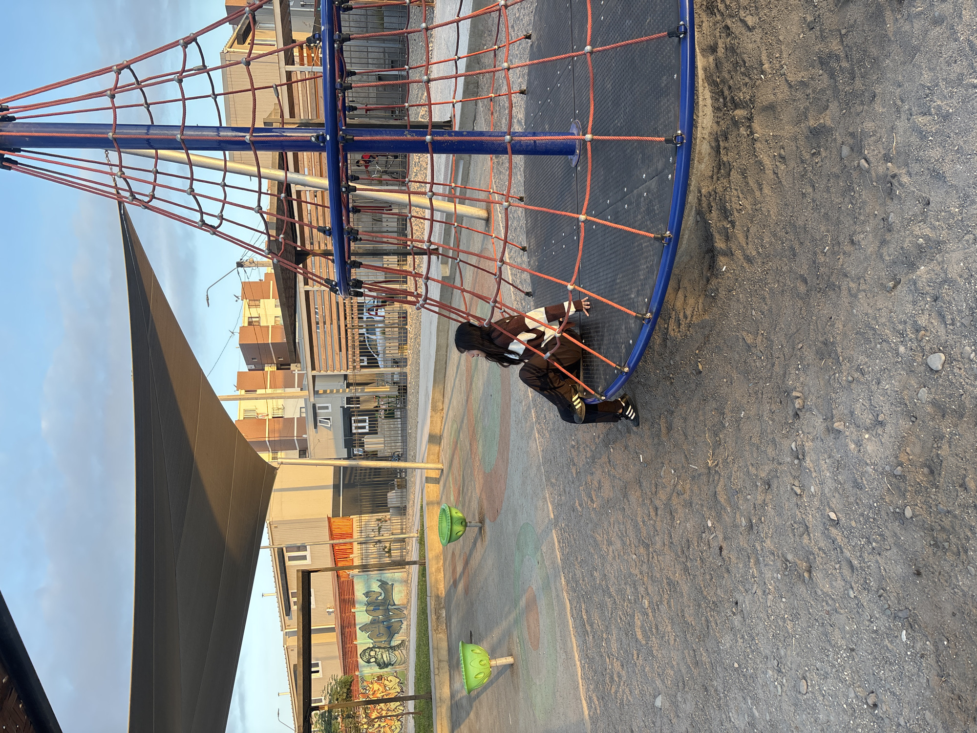 A slightly rotated, medium shot shows a playground scene in Arica, Chile, where a child is climbing on a rope structure. The child, dressed in dark clothing with white stripes on the sleeves, is positioned on the edge of a circular climbing structure with a blue frame and orange rope netting. The structure itself is anchored to the ground and has a central blue pole.
In the background, there are several buildings, including what appears to be prefabricated structures. A large shade sail extends from the left of the frame, casting a shadow over parts of the playground. The ground surface is a mix of dirt and sand. Several green mushroom-shaped objects are scattered across the ground. A painted wall or mural is visible near the buildings. The overall lighting suggests it is late afternoon or early morning.