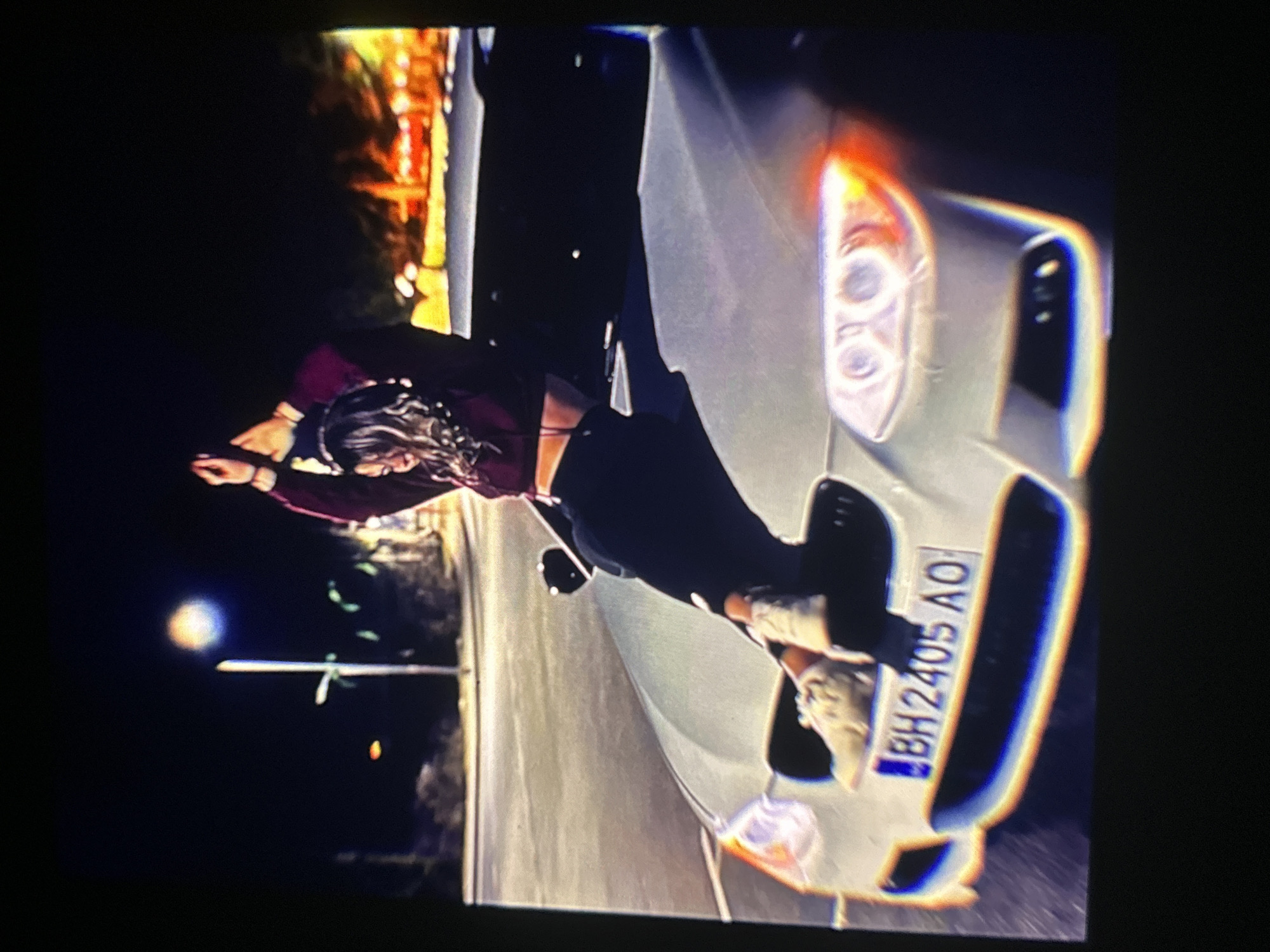 The image depicts a nighttime scene featuring a person lying on the hood of a white car. The individual, seen from behind, has long hair and is wearing a dark top and trousers. Their arms are raised and outstretched, and one foot is casually resting on the car's front bumper.
The car, which appears to be white or light silver, has its headlights illuminated, casting a bright glow that creates some lens flare. A prominent detail is the Bulgarian license plate "BH2405 AO" affixed to the front bumper. The "BH" prefix indicates registration in the Shumen region of Bulgaria, consistent with the provided location context of Bania, Bulgaria.
The setting is outdoors at night, with the background mostly dark except for faint artificial lights, possibly from distant streetlights or buildings, which are out of focus. The scene suggests a casual or staged pose on the vehicle, illuminated primarily by the car's own lights.