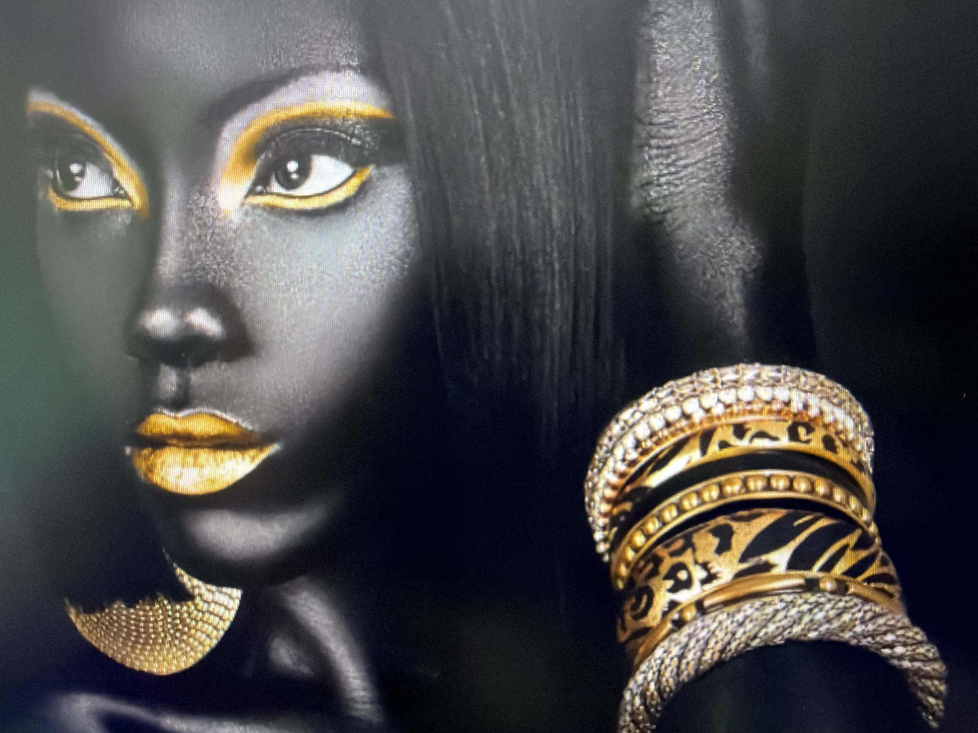 The image displays a close-up, artistic portrait of a person with dark skin. The subject's face is heavily stylized with bold gold eyeshadow that outlines their eyes and is also applied to their lips. The person is adorned with elaborate jewelry. On their left ear, a large, textured gold earring hangs. On their right wrist, they wear a stack of bracelets. The bracelets are a mix of textures and designs, including some with a leopard print pattern and others with intricate beadwork and a braided silver-toned band. The background is dark and indistinct, focusing all attention on the subject and their adornments. The image does not contain any elements that would allow for identification of a specific location, such as buildings, landmarks, or street scenes, and therefore cannot be associated with any city, including Plovdiv, Bulgaria.