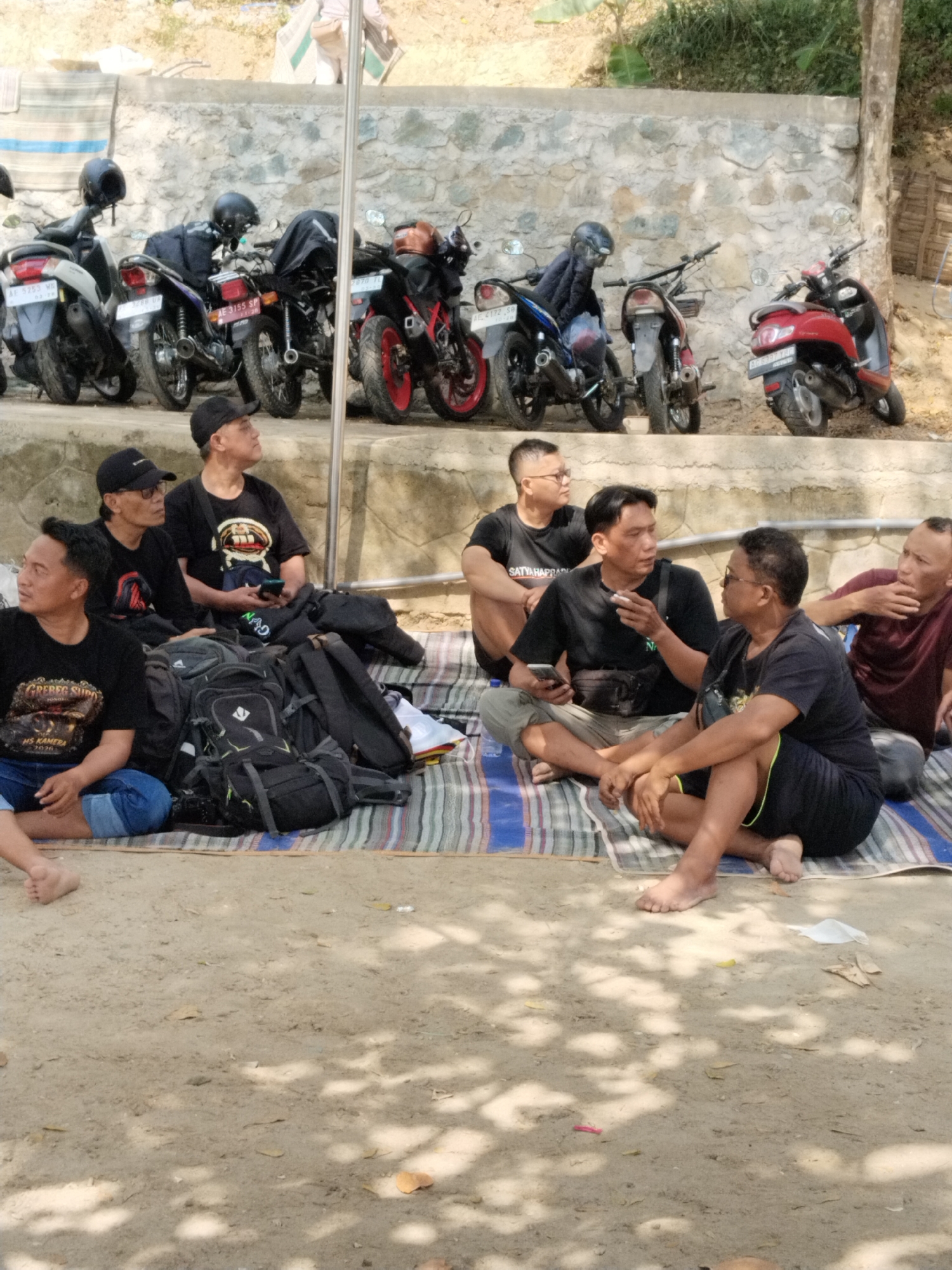 In Prigi, Indonesia, a group of men are gathered outdoors, seated on a striped mat on a sandy area. Behind them, motorcycles are parked in front of a stone wall. The men are dressed casually in t-shirts and shorts. Some are looking at their phones, others are conversing, and one appears to be holding a cigarette. Backpacks are piled near the group. The scene suggests a casual gathering or break.
