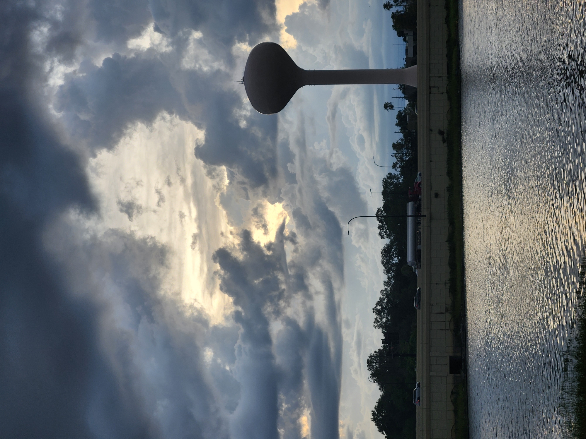 The image depicts a water tower in Altamonte Springs, Florida, United States of America, against a dramatic cloudy sky. The water tower, a prominent white structure with a bulbous top, stands to the right of the frame. Below it, a highway runs horizontally, with cars and a tanker truck visible on the road. The highway is bordered by a concrete barrier and a line of trees. In the foreground, a body of water reflects the cloudy sky and the highway lights. The overall scene is dominated by dark, heavy clouds that obscure much of the sky, with patches of lighter sky and sunlight breaking through in the center.