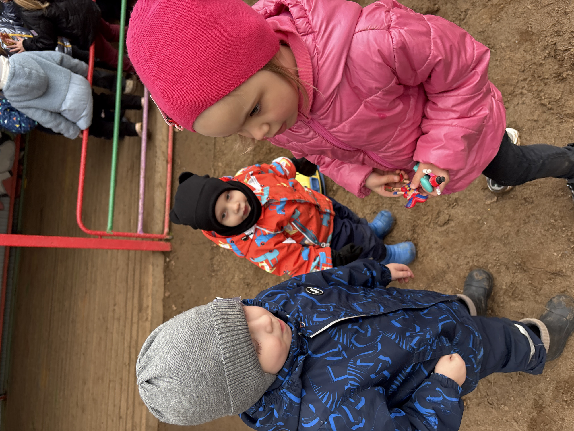 Three young children are the primary subjects in this photograph. One child is wearing a pink hat and pink jacket, and is holding a toy. Another child is wearing a black hat and red patterned jacket. A third child is wearing a grey hat and a blue jacket with blue patterns. The children are outside on a sandy or dirt-covered surface. The setting seems to be near a playground, as suggested by the edge of what appears to be a colorful play structure on the left. It is a day time photo, outdoors, possibly in the city of Chaikovsky, Russia. The location is near a wood surface, likely a playground covering.