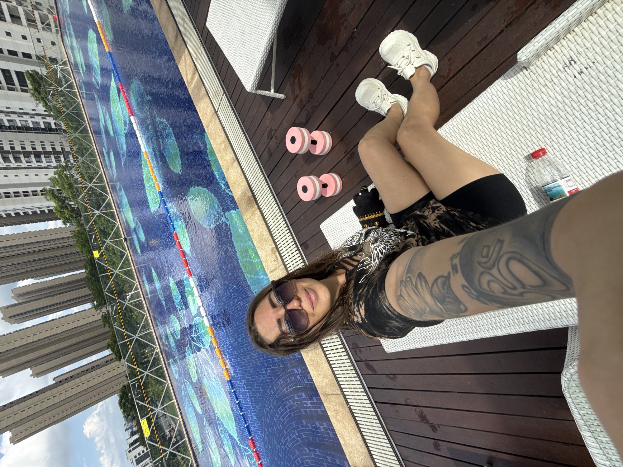 A woman with long brown hair and sunglasses is sitting on a white lounge chair next to a swimming pool. She is wearing a black t-shirt and black shorts. She is holding a phone in her left hand, taking a selfie. Next to her are two pink dumbbells and a water bottle. The swimming pool has lane lines and a decorative pattern on the bottom. In the background, tall apartment buildings are visible. The scene appears to be in the city of Huizhou, China.