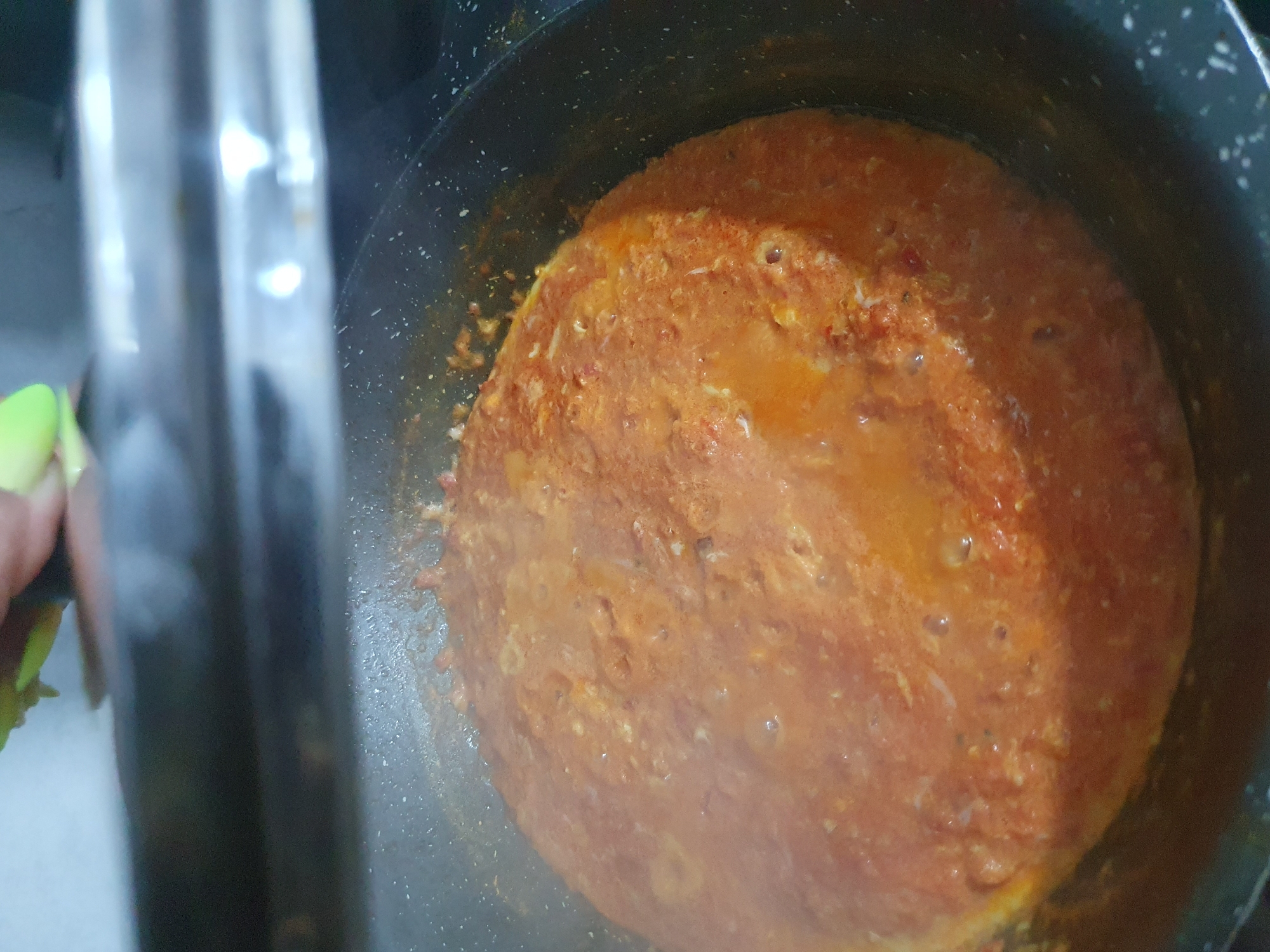An overhead shot captures a close-up view of a bubbling tomato-based sauce cooking in a dark frying pan. The sauce is a vibrant orange-red color and appears to be thick, with small bubbles forming on its surface. The pan has a speckled texture. In the upper portion of the image, a blurred hand is seen holding a bright green object, possibly a piece of food or a utensil. The background is dark and indistinct, suggesting an indoor kitchen setting. The lighting is dim, and the focus is primarily on the food in the pan.