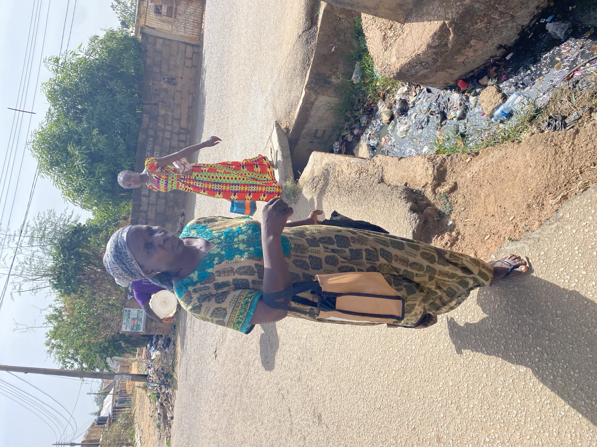 This image depicts an outdoor street scene in Jos, Nigeria, featuring two women. The setting appears to be a residential or semi-urban area, characterized by a paved road, an unpaved shoulder, and simple concrete structures.
The foreground is dominated by an older woman standing on the paved road, facing slightly to her right. She wears a patterned headwrap in white and gray tones, a long-sleeved dress with intricate patterns in green, blue, and beige, and dark flip-flops. She carries a light brown shoulder bag with dark straps. Her expression is neutral.
Behind her, further down the road, stands a younger woman wearing a vibrant, brightly colored dress with geometric patterns in red, orange, green, yellow, and black. She has light-colored hair and is looking towards the camera.
A notable feature of the environment is a concrete drainage ditch or gutter running parallel to the road. This gutter is filled with murky water and a significant amount of trash, including plastic bottles, bags, and other debris, indicating poor waste management. The ground bordering the gutter is rough and unpaved, with some sparse patches of grass.
In the background, there are green trees and bushes, suggesting a lush, sunny environment. Some basic concrete walls and parts of buildings are visible. Overhead, power lines can be seen against a bright sky, indicating it is daytime with clear weather. The shadows cast by the subjects suggest the sun is high, likely mid-morning to early afternoon.
A white and green sign, possibly an informational or public notice board, is visible in the background near a tree, but its text is illegible. The overall scene conveys a slice of daily life in a bustling community.