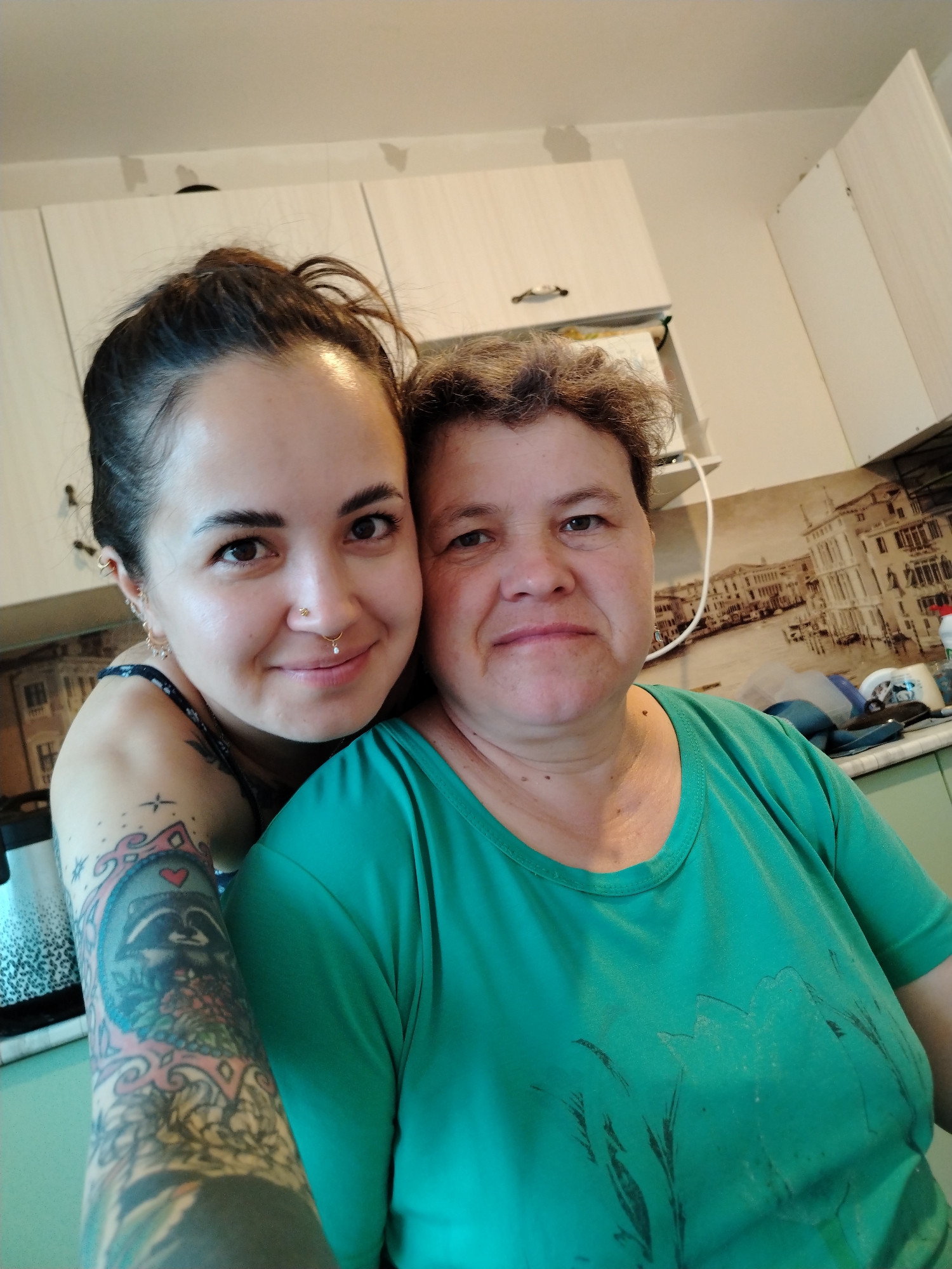 In a dimly lit kitchen, two women, likely mother and daughter, pose for a selfie. The younger woman, on the left, leans forward, her face partially obscured by her mother's head. She has dark hair pulled back in a messy bun and a nose ring. Her arm, adorned with a colorful tattoo of a raccoon, is visible. Her mother, on the right, has short, curly brown hair and wears a bright green t-shirt. They stand in front of kitchen cabinets and a backsplash decorated with a cityscape. The overall lighting suggests it's indoors, possibly in a residential kitchen. No specific location within Noviy, Russia is identifiable from the image.