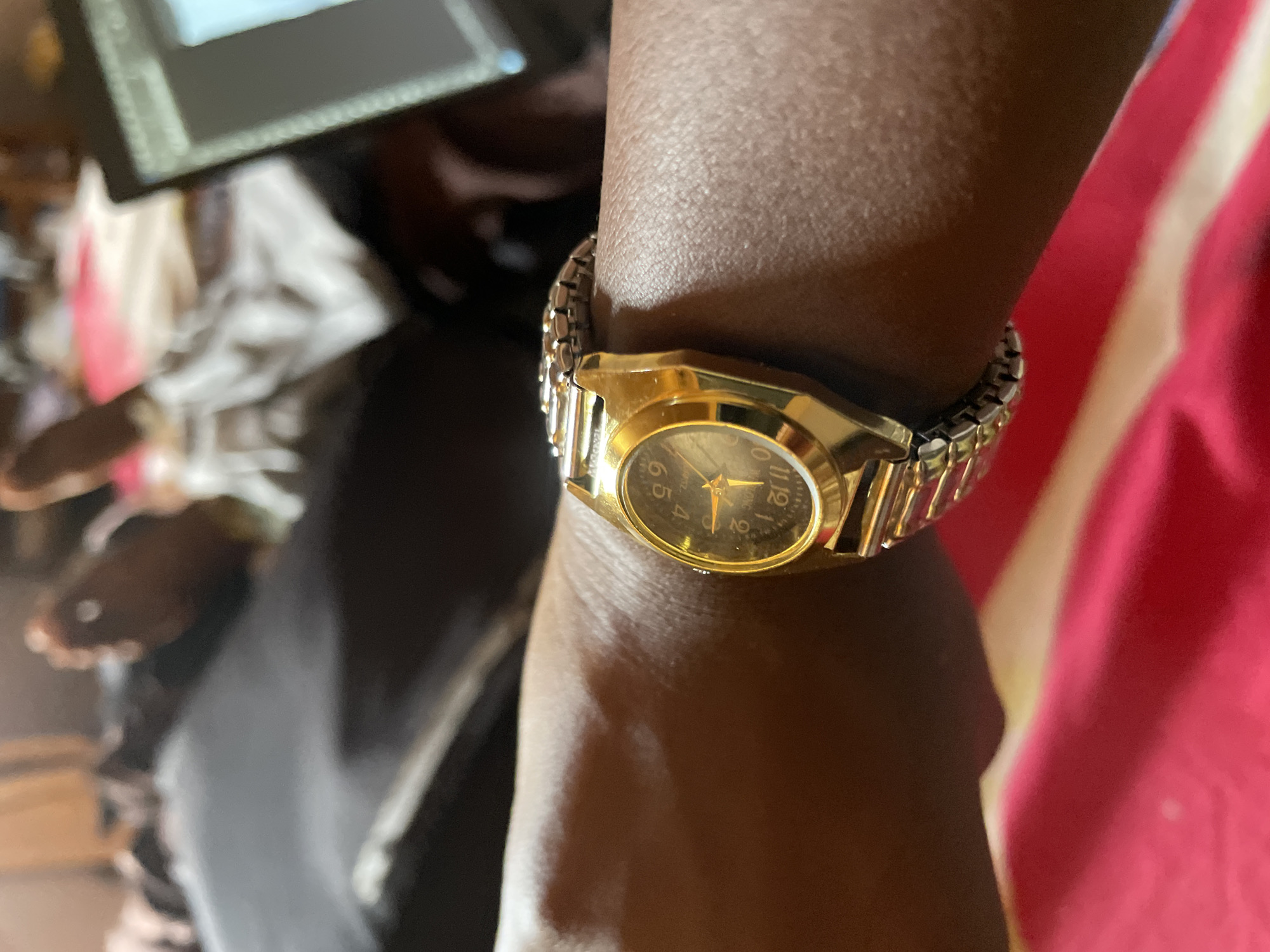 A close-up image shows a dark-skinned person's wrist adorned with a gold-toned wristwatch with a metal band. The watch face features numeric indices and gold hands. The background is slightly blurred, revealing other objects including a screen and a piece of red and white fabric. The location is unverifiable as Jos, Nigeria.