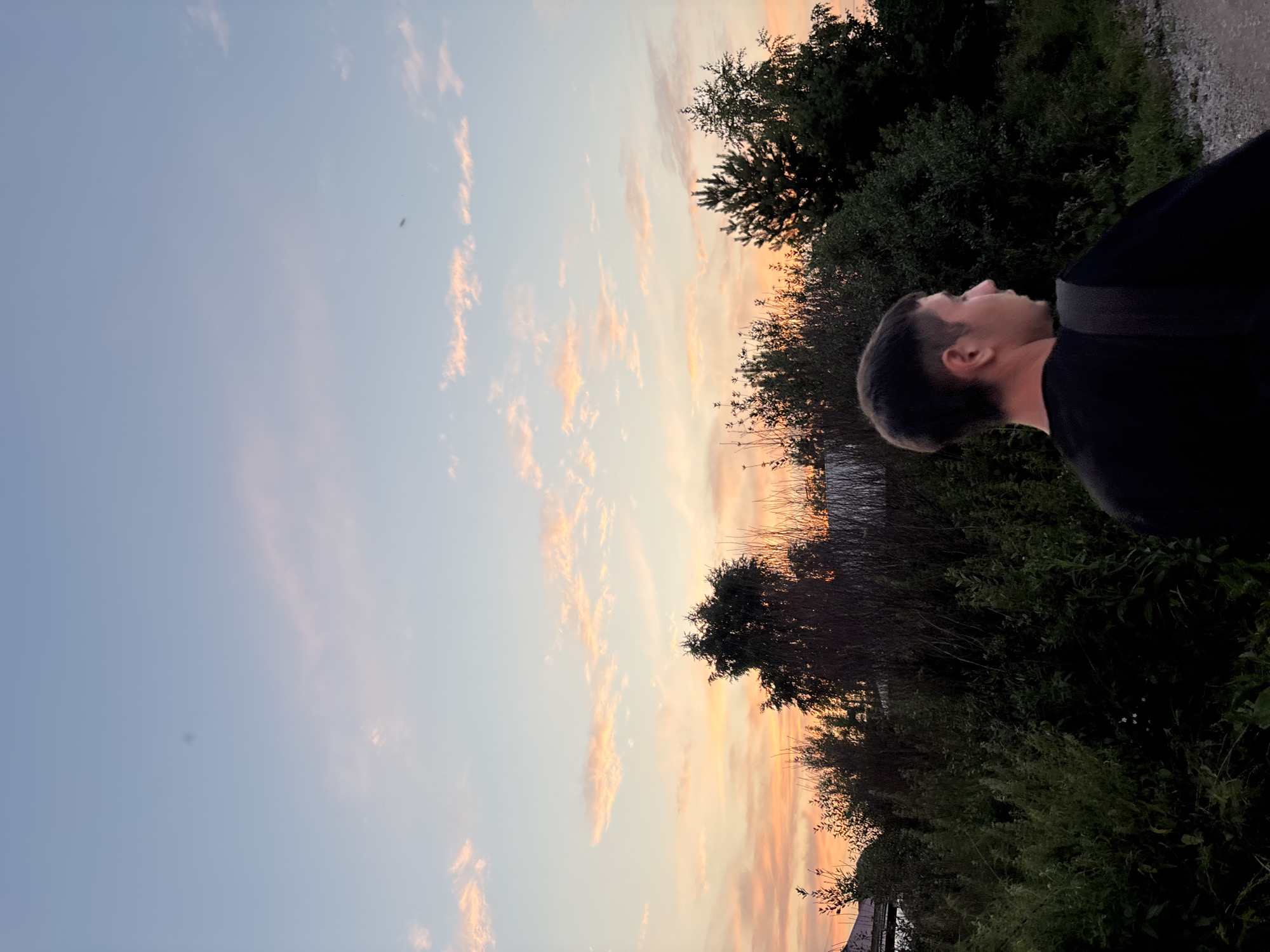 A young Caucasian male with short, dark hair is captured from the side, looking away from the camera towards a sunset over Vishnevka, Russia. He is wearing a black t-shirt and a dark backpack slung over his right shoulder. The background features a silhouette of trees and vegetation against a sky filled with soft pink and orange clouds. The scene is set in a rural area, with a dirt path visible in the foreground.