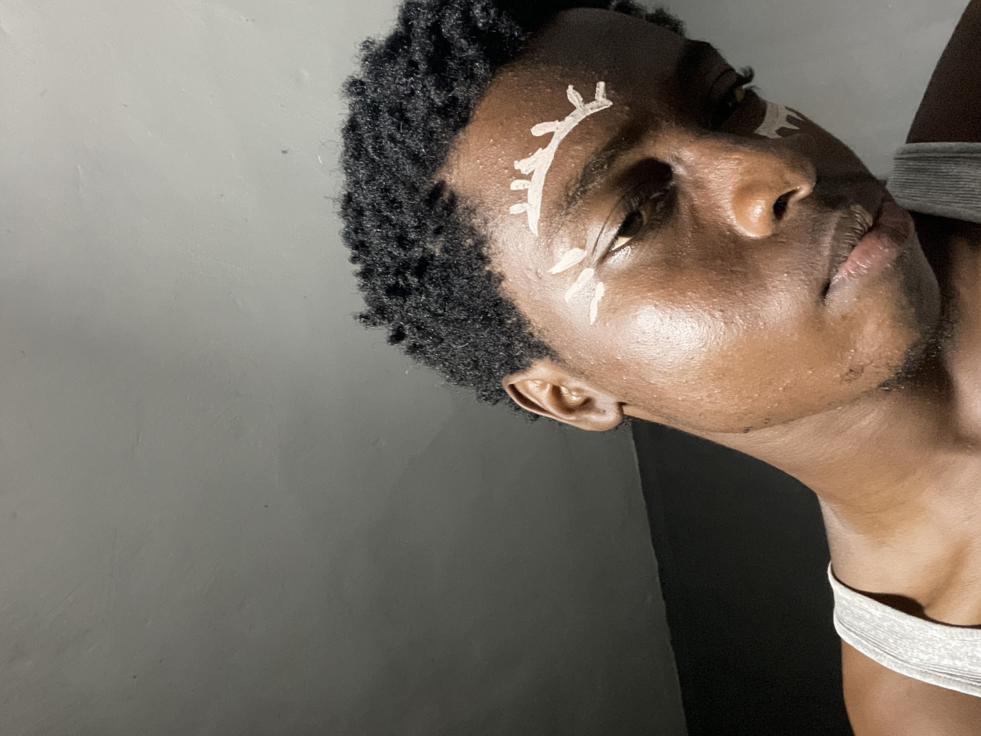 The image shows a close-up portrait of a young Black person with short, dark, curly hair and dark skin. They are wearing a grey tank top. White face paint is applied in a decorative pattern around their eyes and on their forehead, resembling stylized eyebrows and possibly markings. The background appears to be a grey wall and a darker area, possibly a ceiling or a different wall. The lighting suggests an indoor setting. There is no indication of the city of Nkpor, Nigeria, within the image itself.