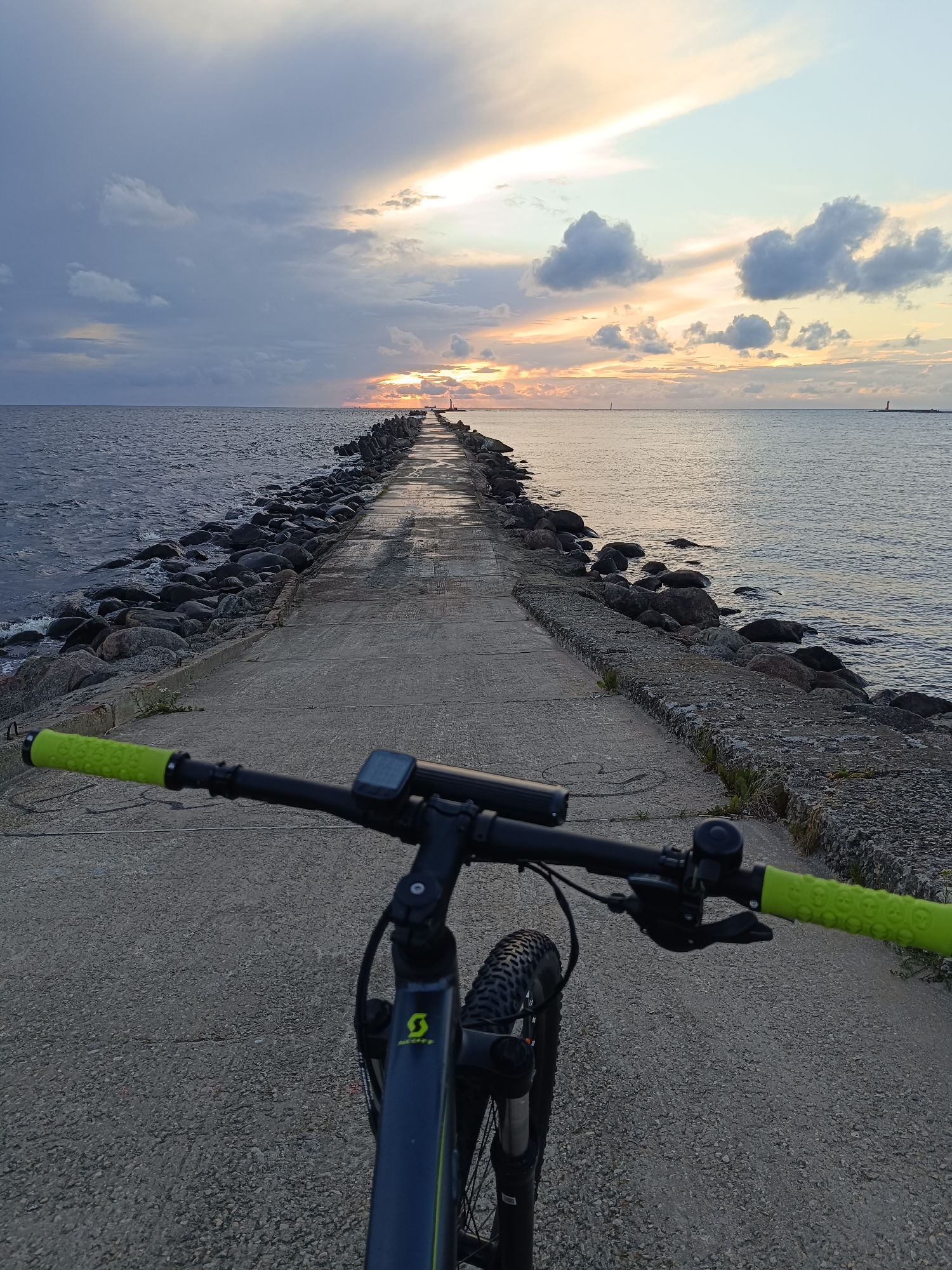 The image shows a first-person perspective of a bicycle's handlebars and front wheel, positioned on a concrete pier. The pier extends into a calm body of water, likely the Baltic Sea, with a rocky breakwater on the left. In the distance, a lighthouse stands at the end of the pier. The sky is filled with dramatic clouds and a sunset with streaks of orange and yellow. This scene is consistent with the Daugava River mouth or the Baltic Sea coast near the city of Riga, Latvia, possibly near the Andrejosta or Vakarbuļļi areas, known for their piers and coastal landscapes. The bicycle appears to be an adult mountain bike, with neon green grips and a black frame. The overall atmosphere is serene and picturesque, capturing a moment of outdoor activity at dusk.