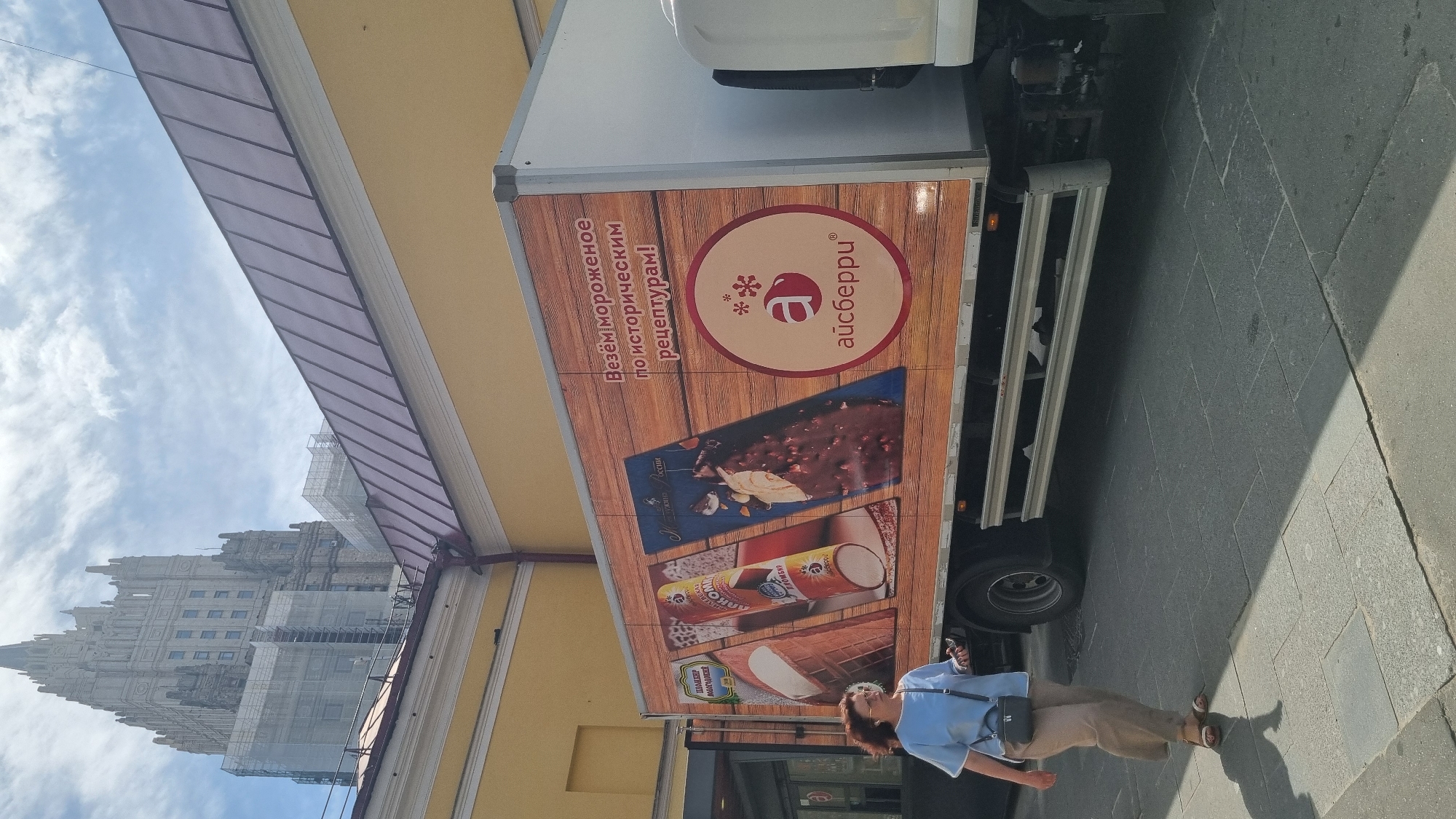 A woman wearing a light blue short-sleeved shirt and beige pants walks on a stone-paved sidewalk next to a delivery truck. The truck has a wood-grain exterior on its side, displaying advertisements for ice cream. The advertisements feature images of ice cream cones and bars, with text in Russian that translates to "We deliver ice cream according to historical recipes!" and the brand name "Aisberri" with a logo featuring a stylized red 'a' and snowflakes. In the background, a tall, imposing building, likely a Stalinist skyscraper, is visible, with scaffolding on its upper floors, suggesting ongoing construction or renovation. The sky is partly cloudy. The scene appears to be in an urban environment.